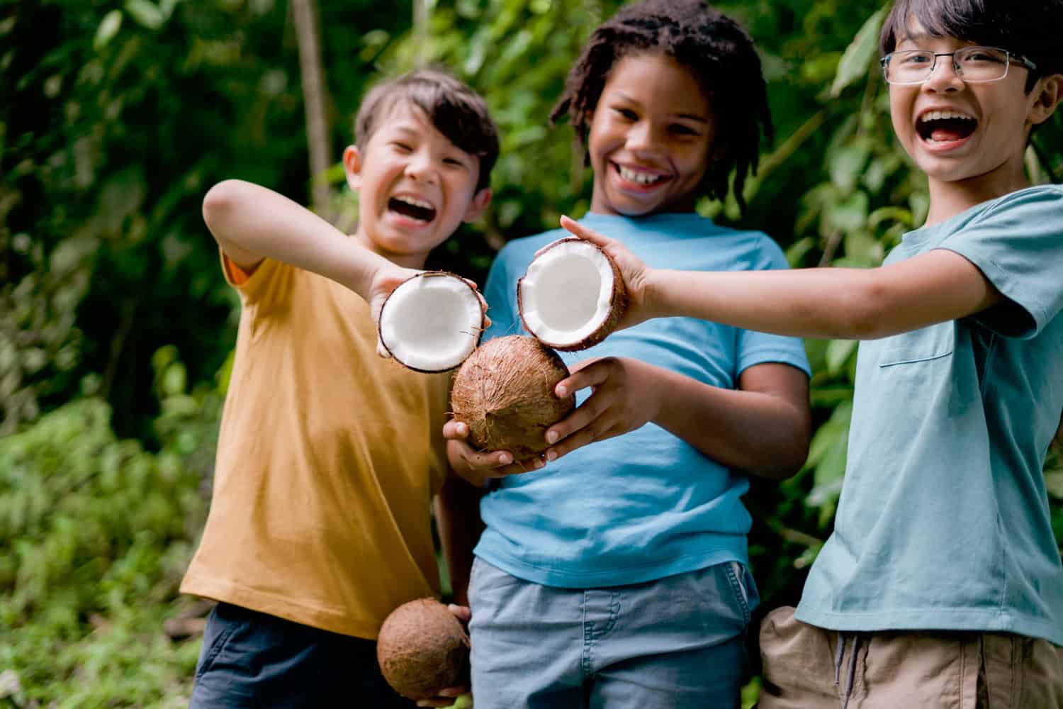 JT-Family-Travel-12 Three children smiling outdoors hold and display opened coconuts, with green foliage in the background.