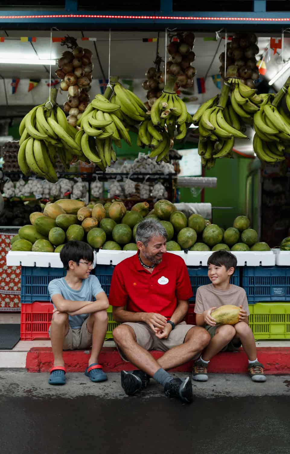 JT-Family-Travel-13 An older man sits on a curb with two boys in front of a fruit stand displaying bananas and other produce. The boys appear to be holding a melon.