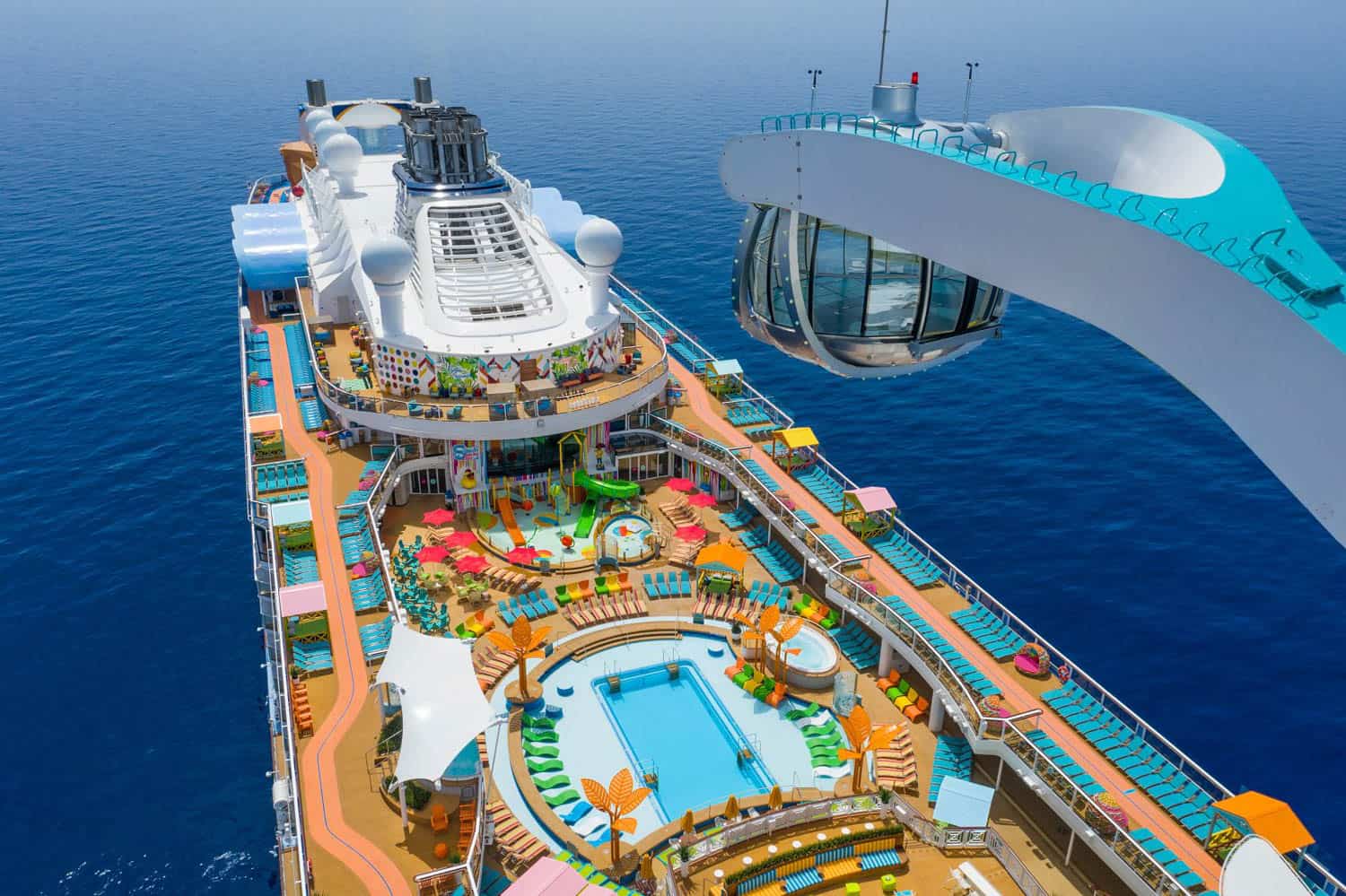 Aerial view of a large cruise ship with pools, lounge chairs, a glass observation capsule, and multiple decks, surrounded by calm blue ocean.