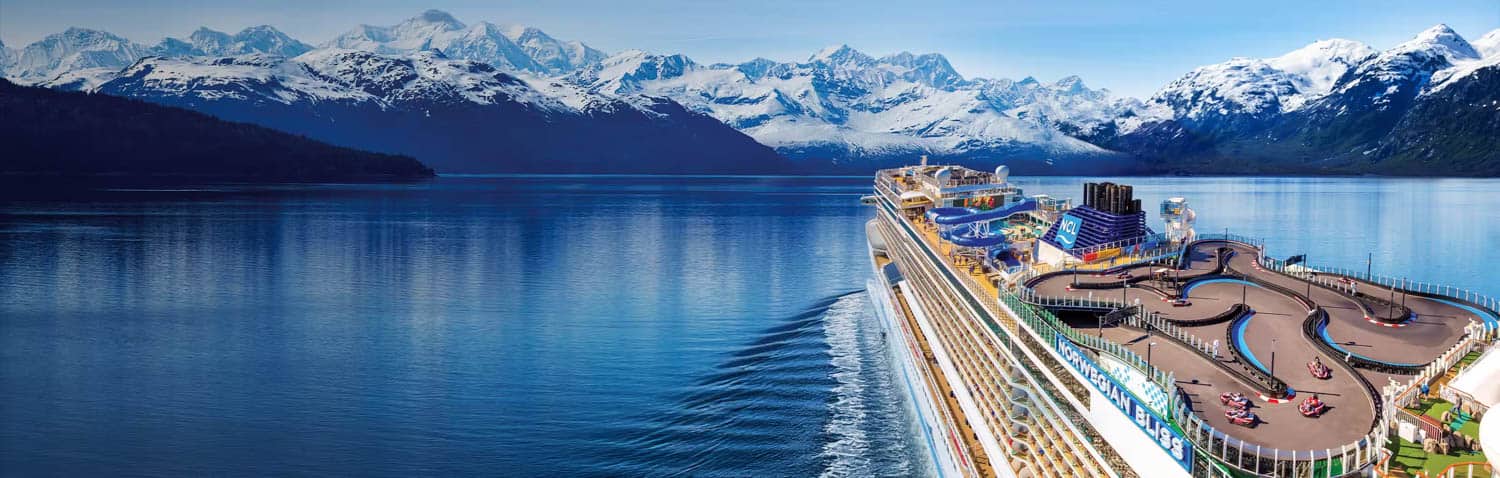 A large cruise ship in Alaska, with a go-kart track on its deck sails through calm blue water, surrounded by snow-capped mountains under a clear sky.