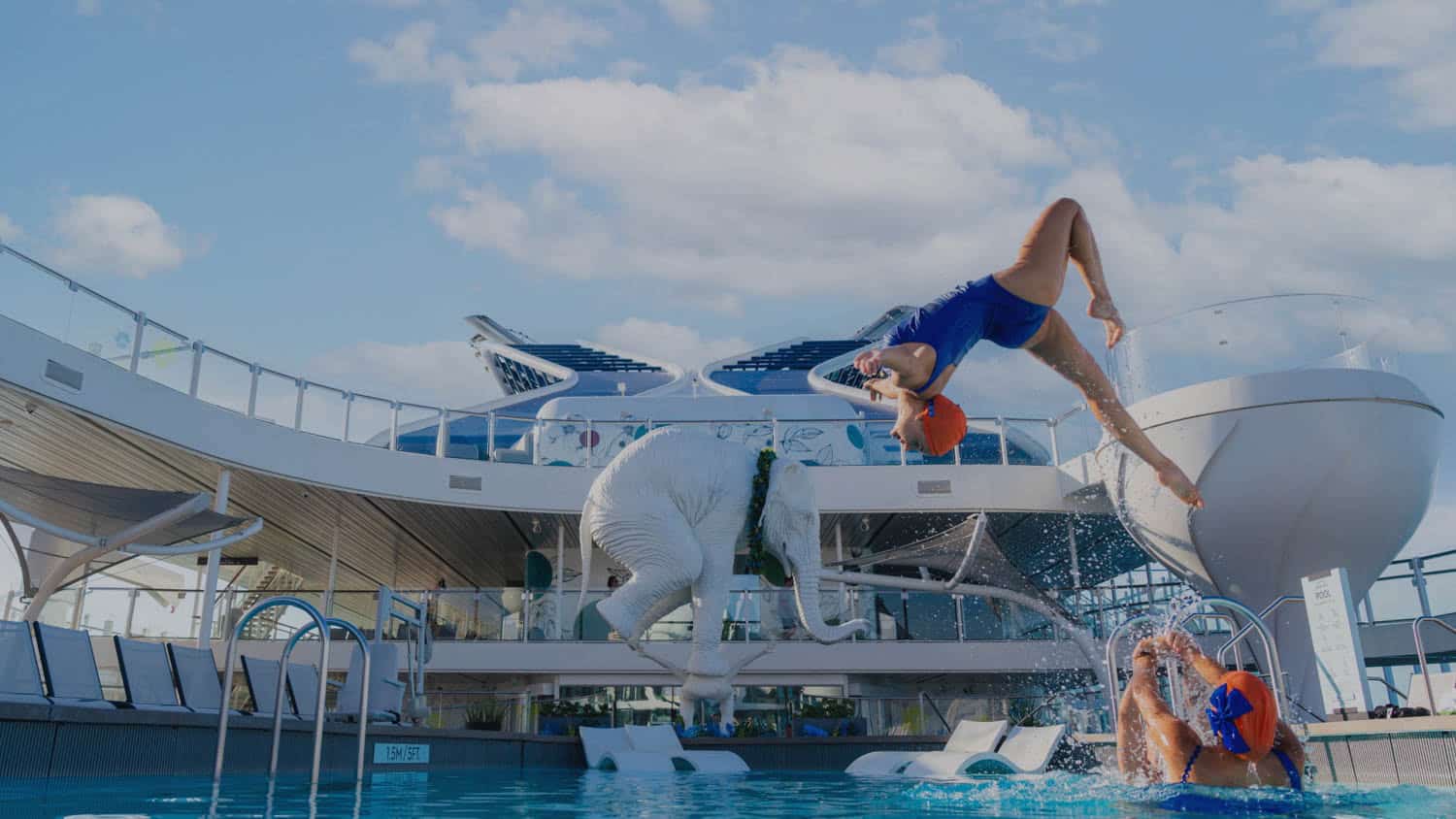 A woman in blue swimwear flips backward into a cruise ship pool while another woman and a white pool float shaped like an elephant are in the water.