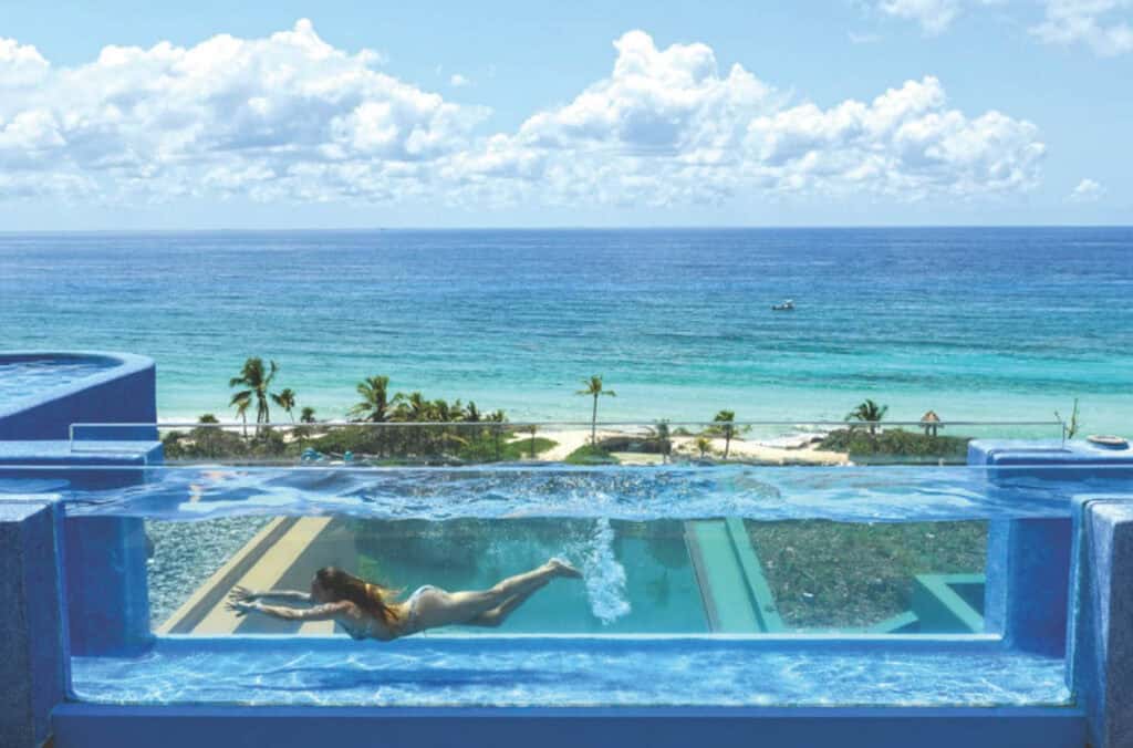 A woman swims in a rooftop infinity pool overlooking a beach, palm trees, and the ocean under a partly cloudy sky.