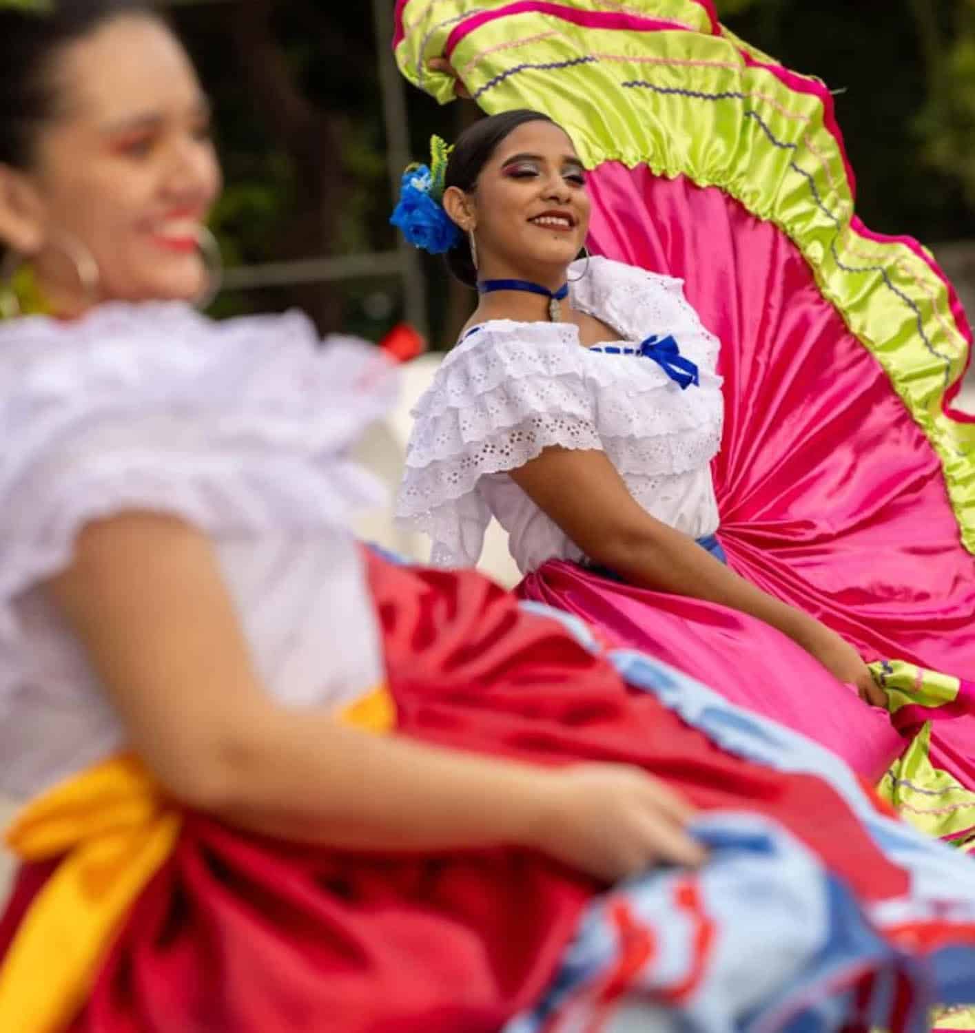 Two women in traditional Mexican dresses perform a folk dance, one in focus wearing a pink and green skirt, the other blurred in the foreground.