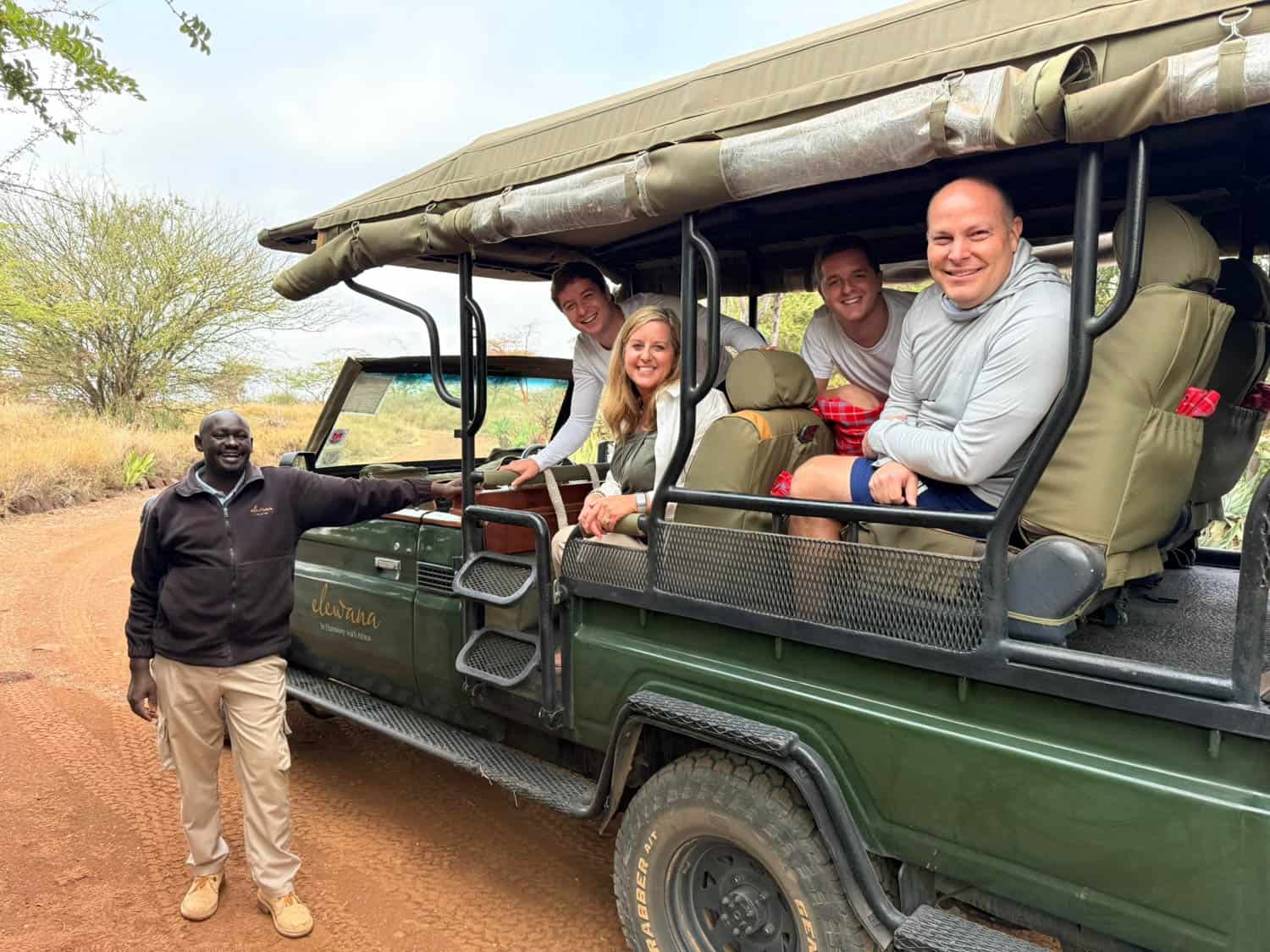 Five people on an African safari pose by a vehicle on a dirt road. Four are seated inside while one stands beside the vehicle in outdoor clothing.