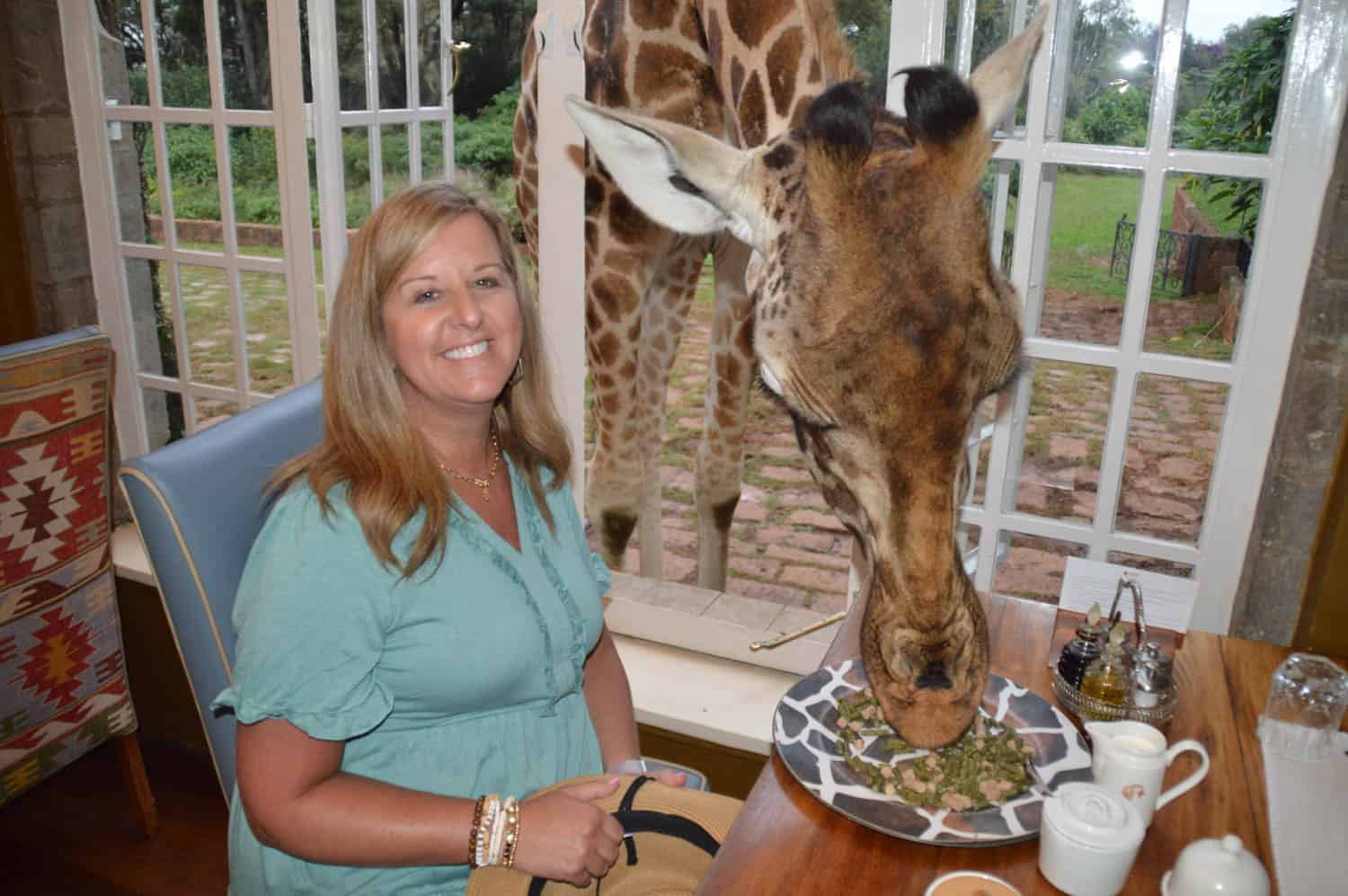 A woman on an African safari sits at a table smiling while a giraffe eats from a plate through an open window behind her.