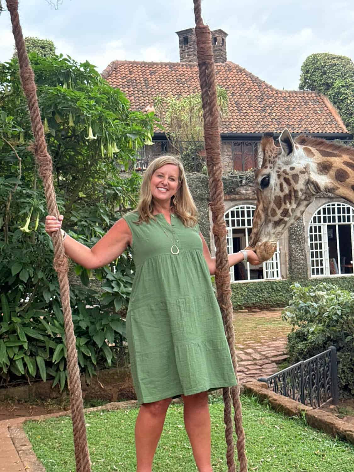 A woman in a green dress stands on a swing, smiling and feeding a giraffe in a garden with a brick house in the background.