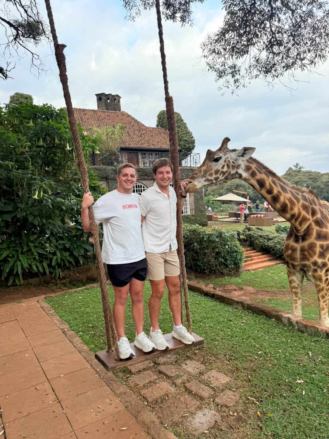 Two men stand on a wooden swing while a giraffe stands nearby. There is greenery, a brick path, and a large house in the background.