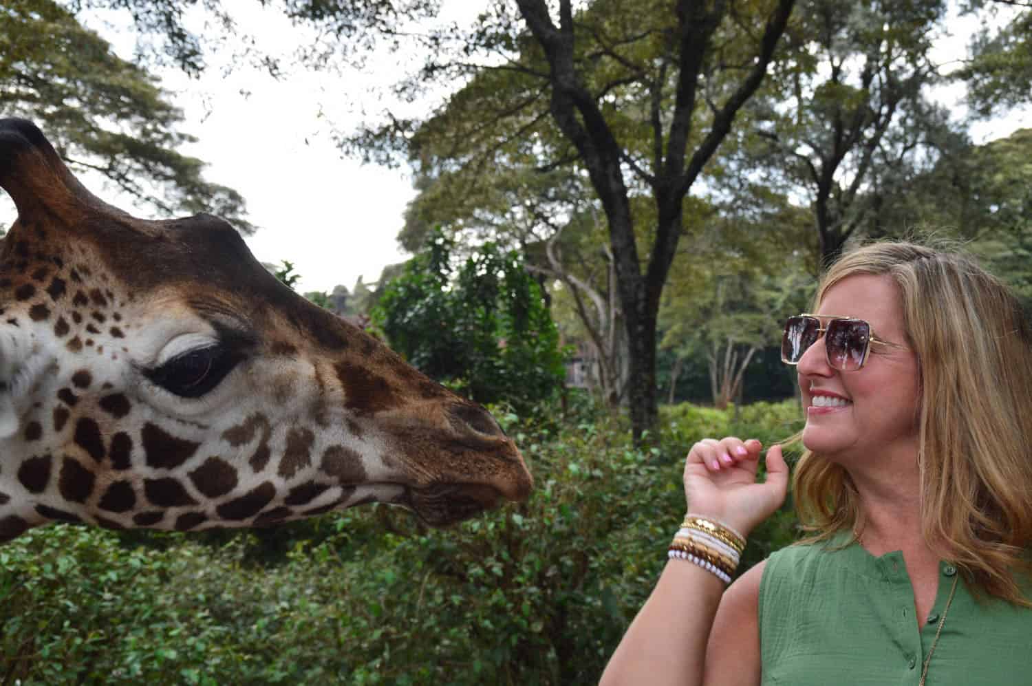 A woman on an African safari wearing sunglasses and a green top, smiles while standing close to a giraffe.