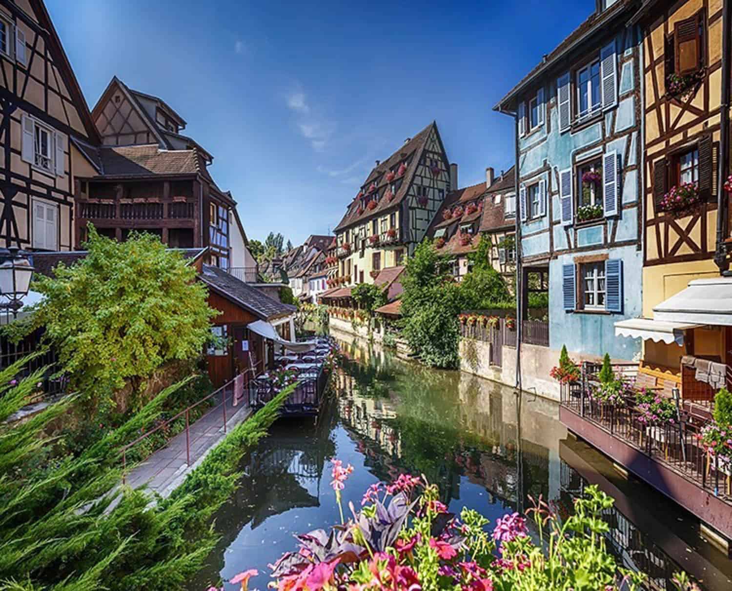 Anna-Swenson-07 A canal lined with colorful half-timbered houses and flowers runs through a picturesque European village under a clear blue sky.