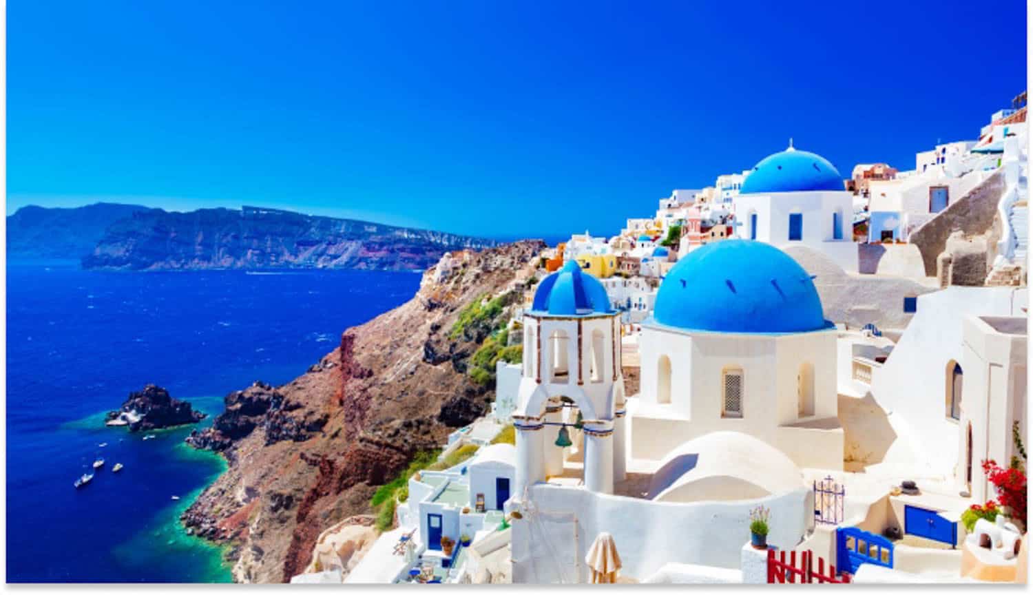 Anna-Swenson-10 White buildings with blue domes overlook the deep blue sea and rugged coastline of Santorini, Greece, under a clear, sunny sky.