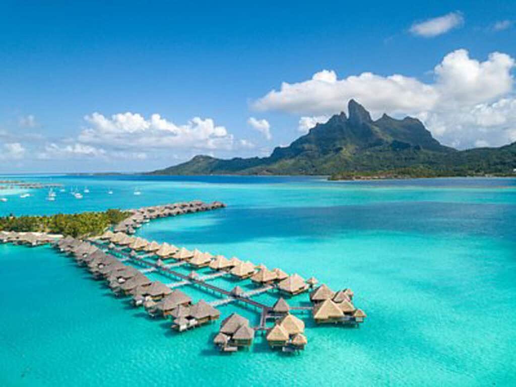 Overwater bungalows stretch into a turquoise lagoon with a green mountain and scattered clouds in the background.