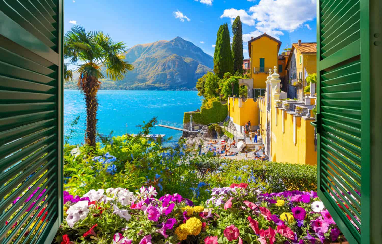 Anna-Swenson-17 View through green window shutters of colorful flowers, yellow buildings, and Lake Como with mountains in the background under a bright blue sky.
