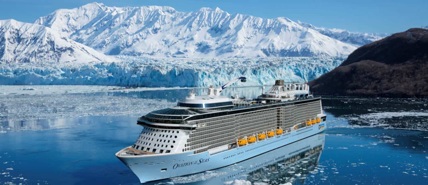 Anna-Swenson-18 A large cruise ship sails through icy waters with a backdrop of snow-covered mountains and a glacier under a clear blue sky.