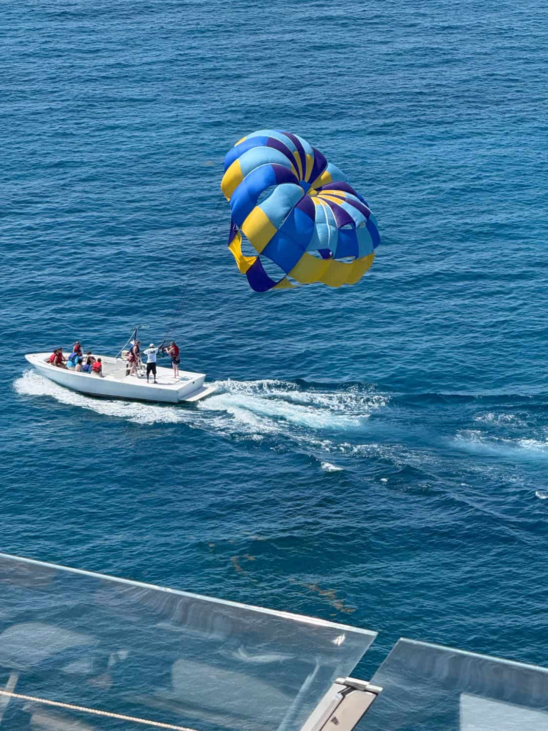 A speedboat pulls a person parasailing with a blue and yellow parachute over the ocean, while people watch from the boat. Glass panels are in the foreground.