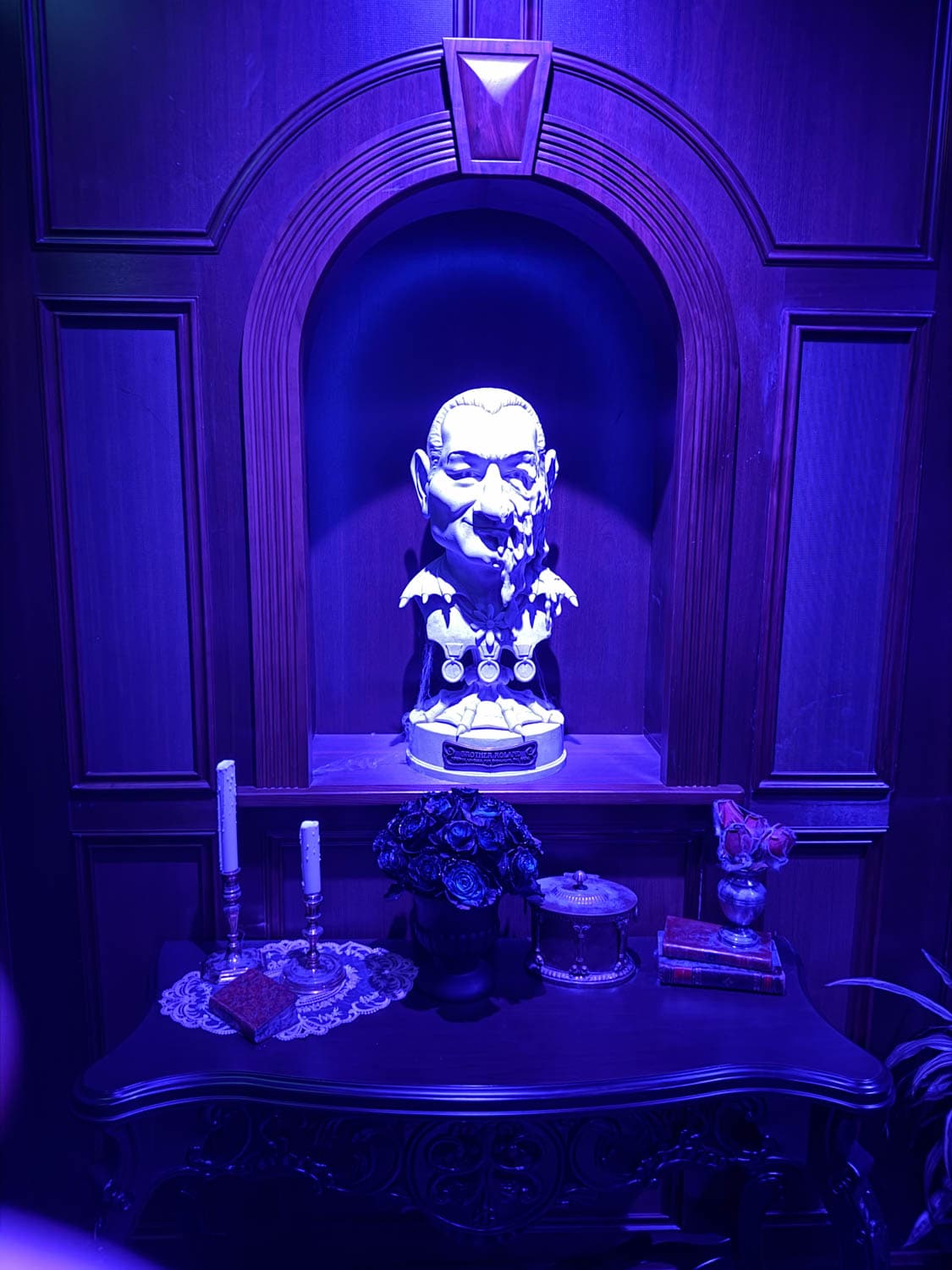A bust of a man with medals displayed in an alcove, illuminated by blue light, with candles, flowers, and books on a table below.