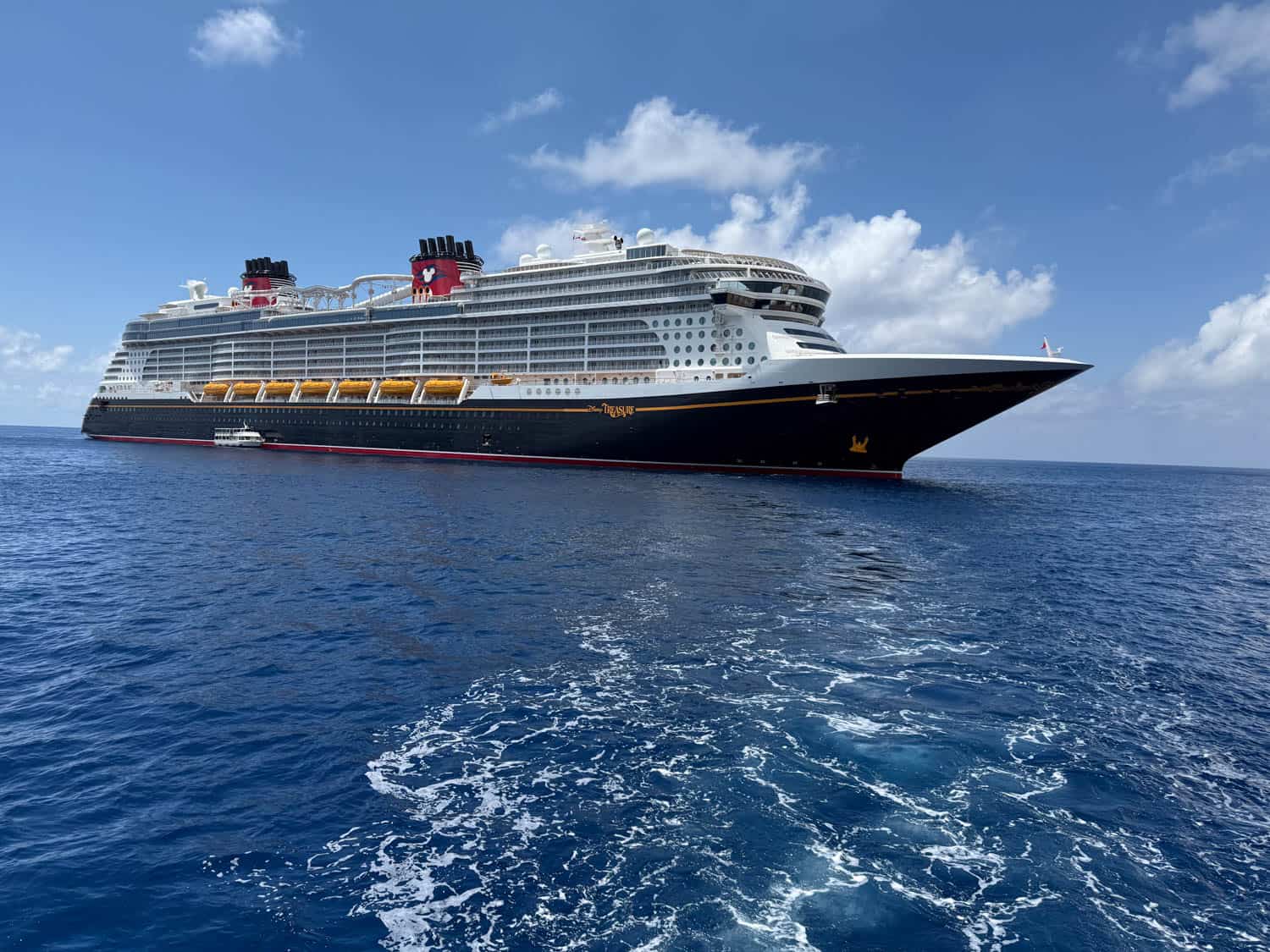 A large cruise ship with red and black funnels sailing on calm blue ocean water under a partly cloudy sky.
