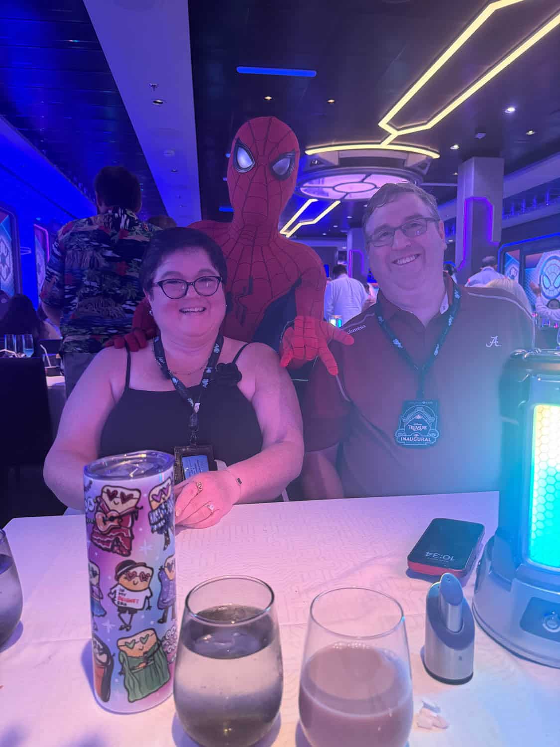Two people sit at a table in a themed restaurant, smiling for a photo with a man dressed as Spider-Man behind them. Glasses and drinks are on the table.