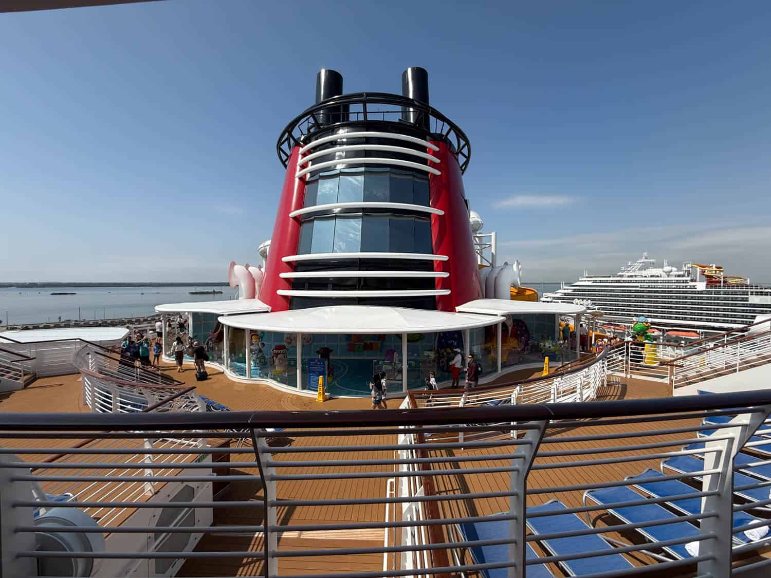 A view of a cruise ship deck with lounge chairs, railings, and a large red and black funnel structure in the center. Another cruise ship is in the background.