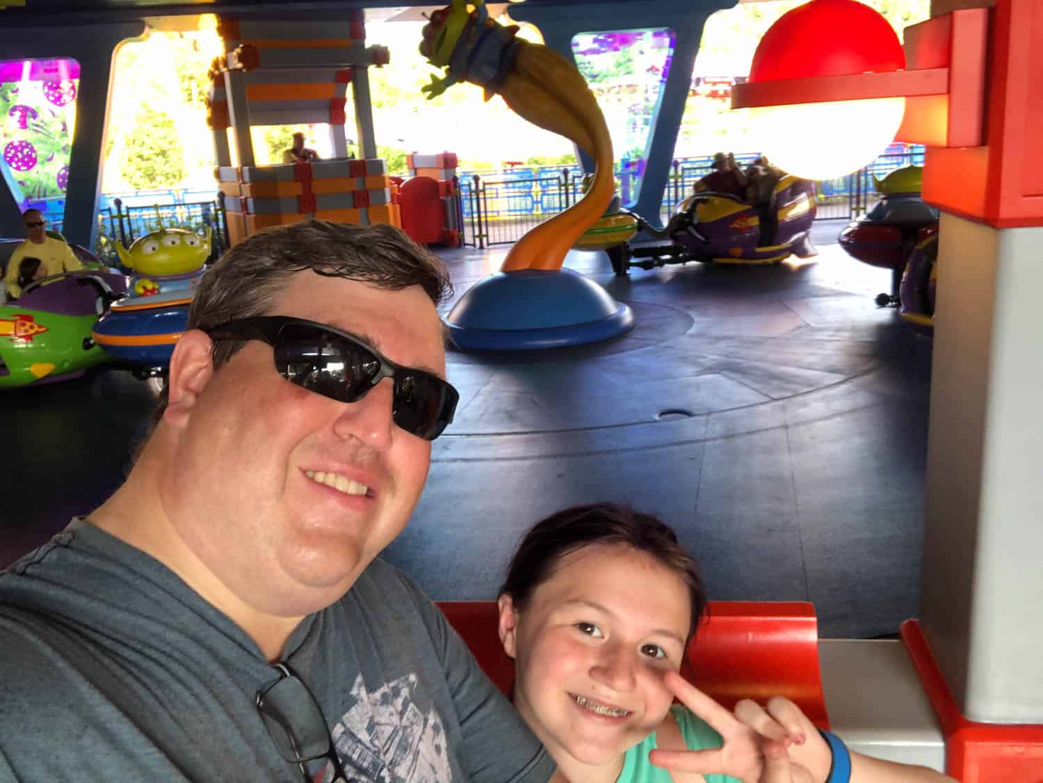A man wearing sunglasses and a girl smiling pose for a selfie at an amusement park ride themed with colorful alien spaceships.