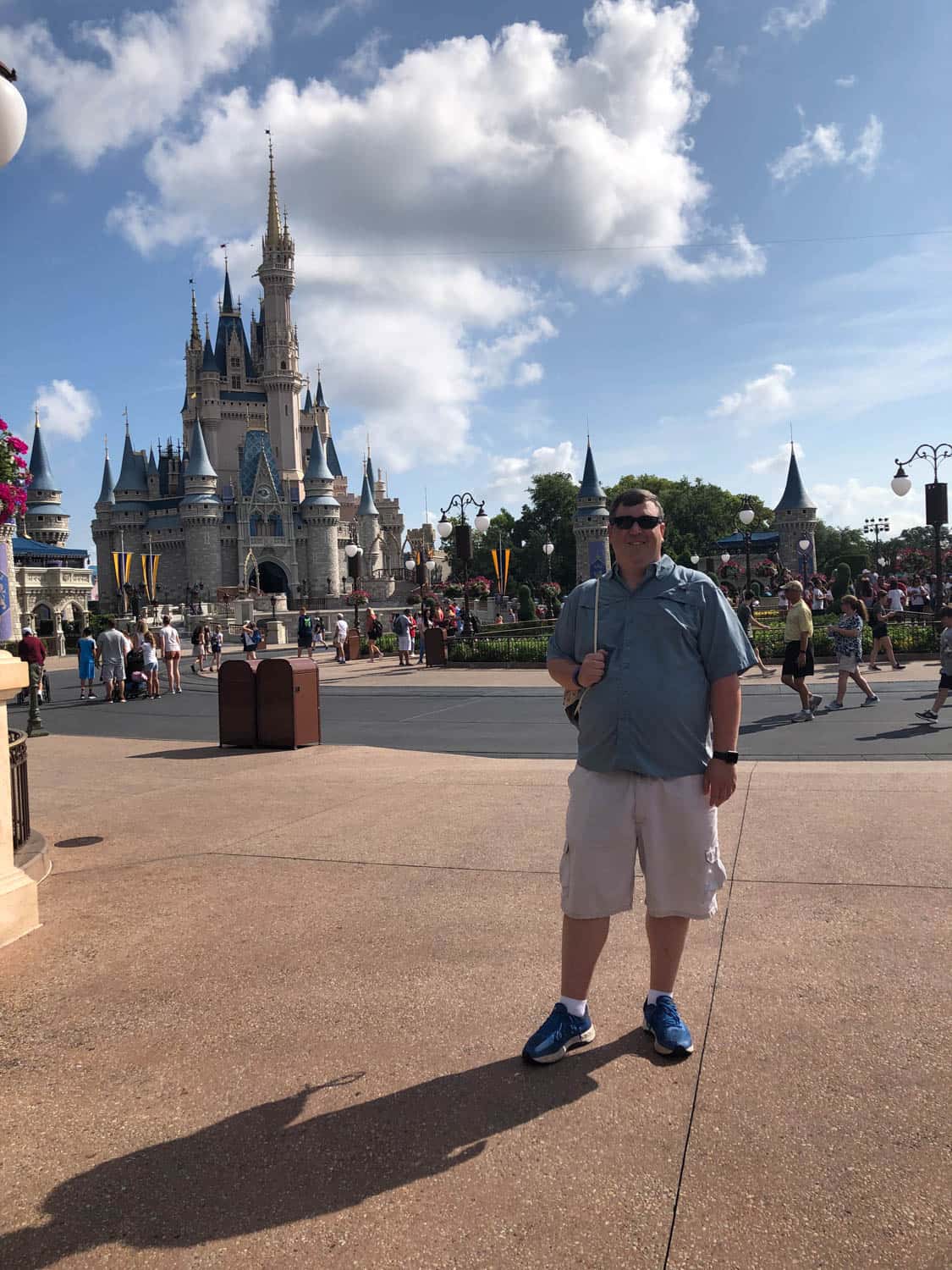 A man in sunglasses stands in front of Cinderella Castle at a Disney theme park on a sunny day.
