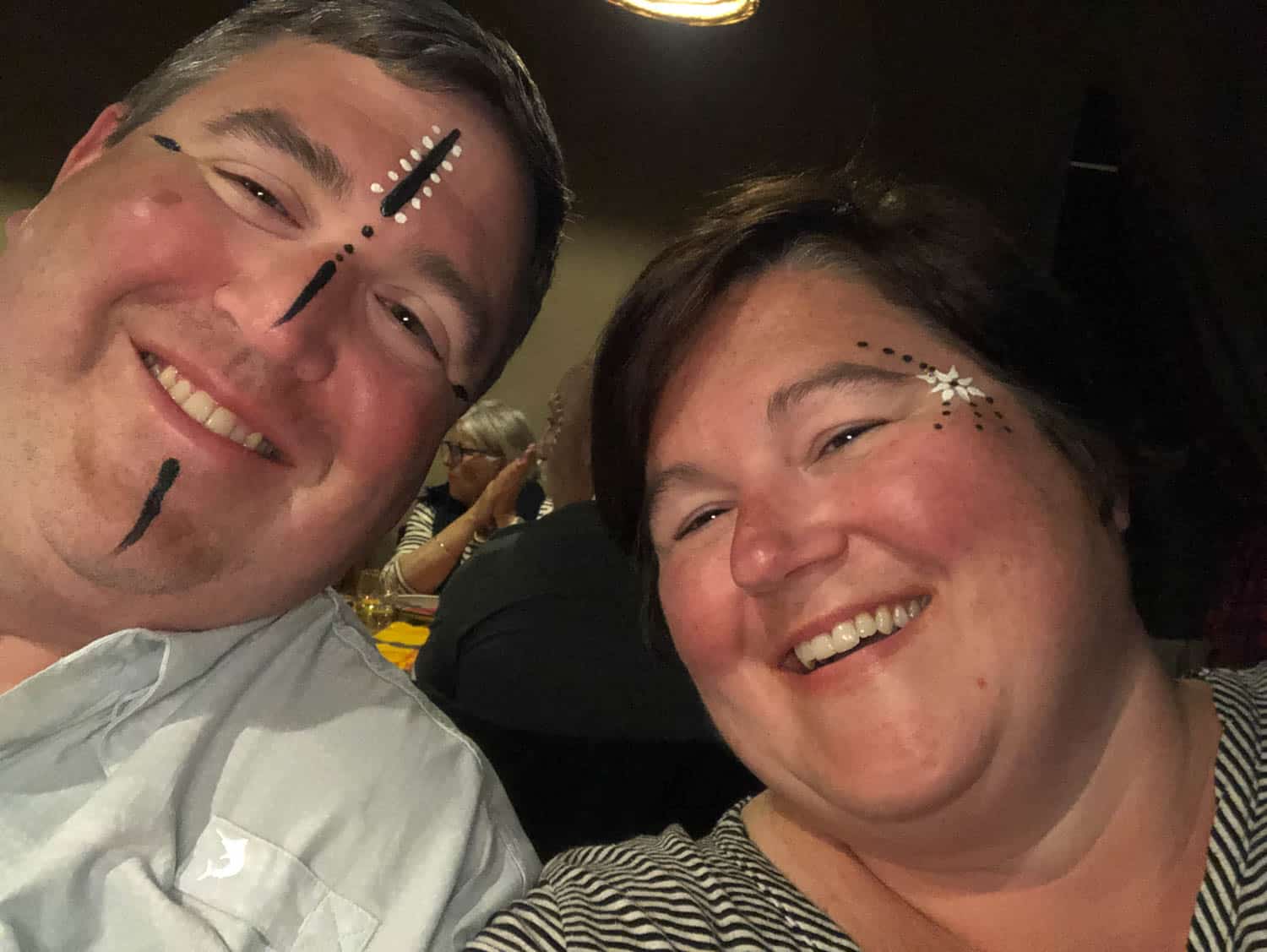 A man and a woman smiling, both with face paint featuring black lines and white dots on their faces, sitting indoors.