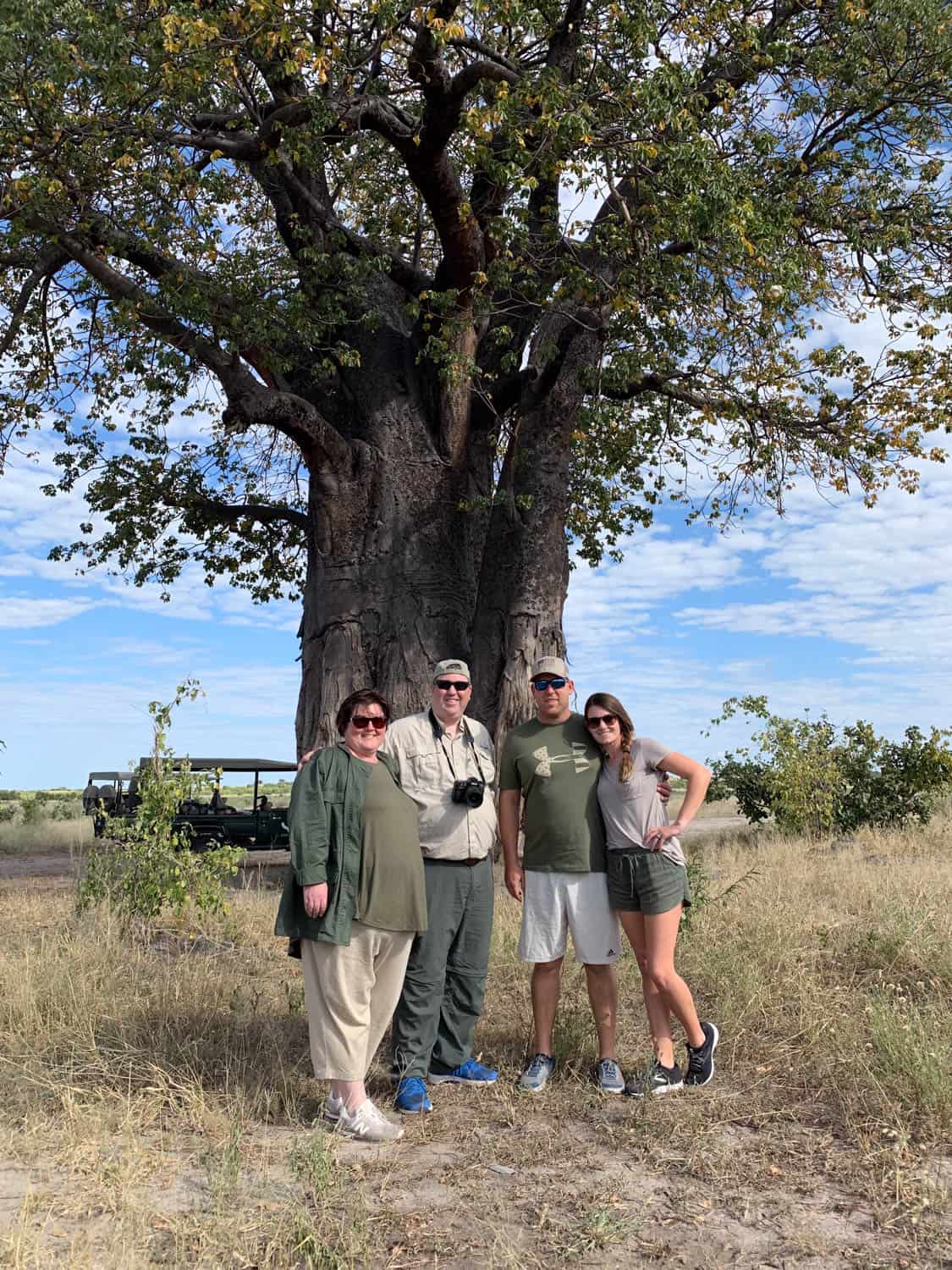 Four people stand in front of a large tree in a grassy area, with a safari vehicle in the background under a partly cloudy sky in Africa.