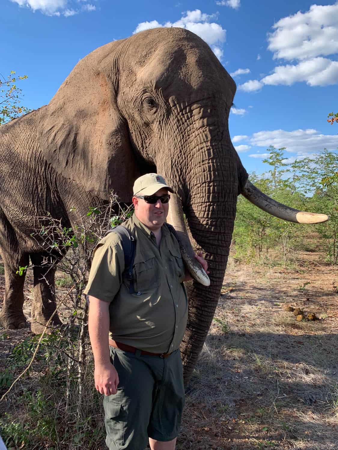 A man in safari clothing stands in front of a large elephant in a natural, wooded area under a blue sky with scattered clouds in Africa.