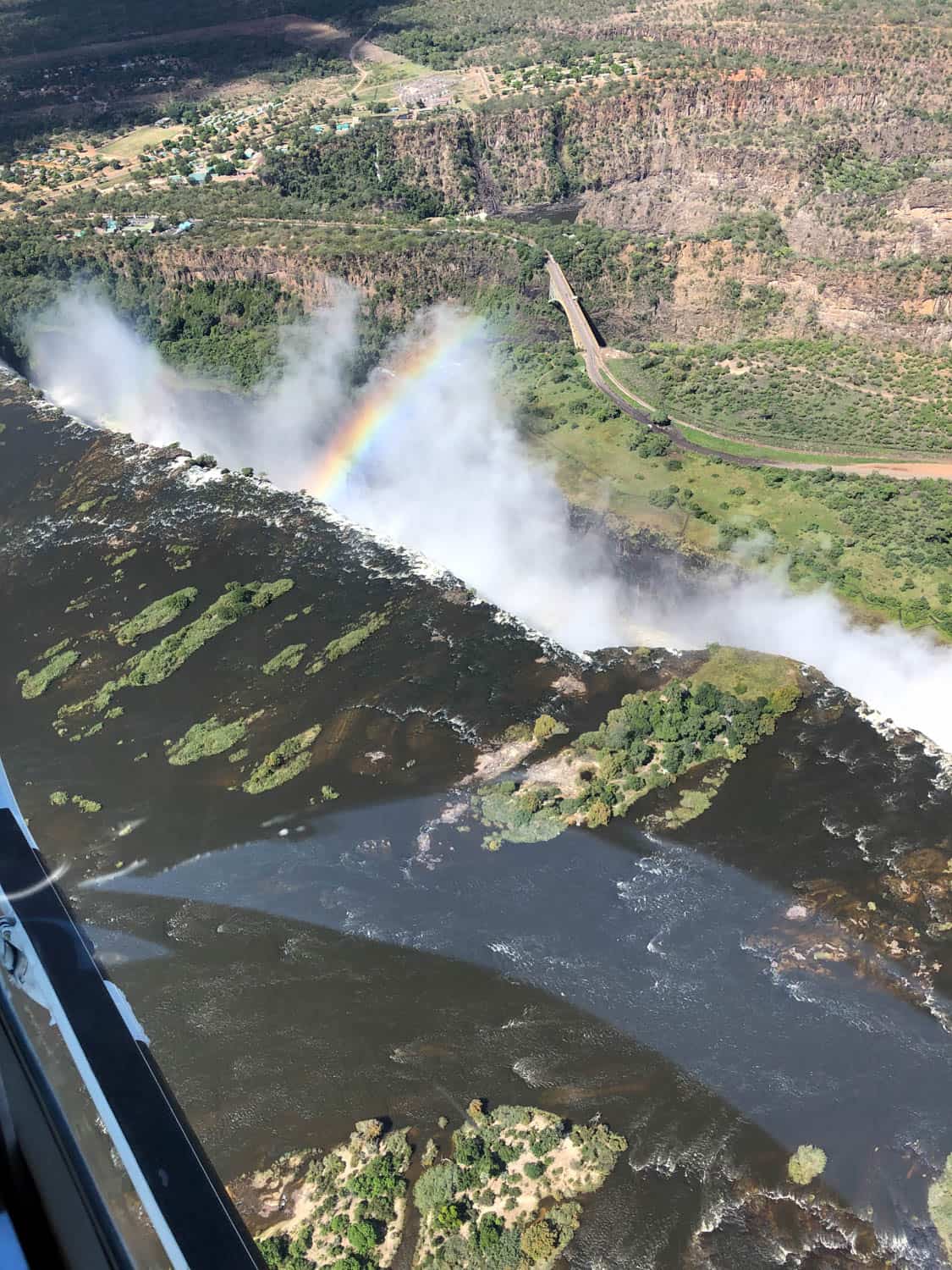 Aerial view of a large waterfall creating mist, with a rainbow visible above the falls and a winding road along the cliff in the background.
