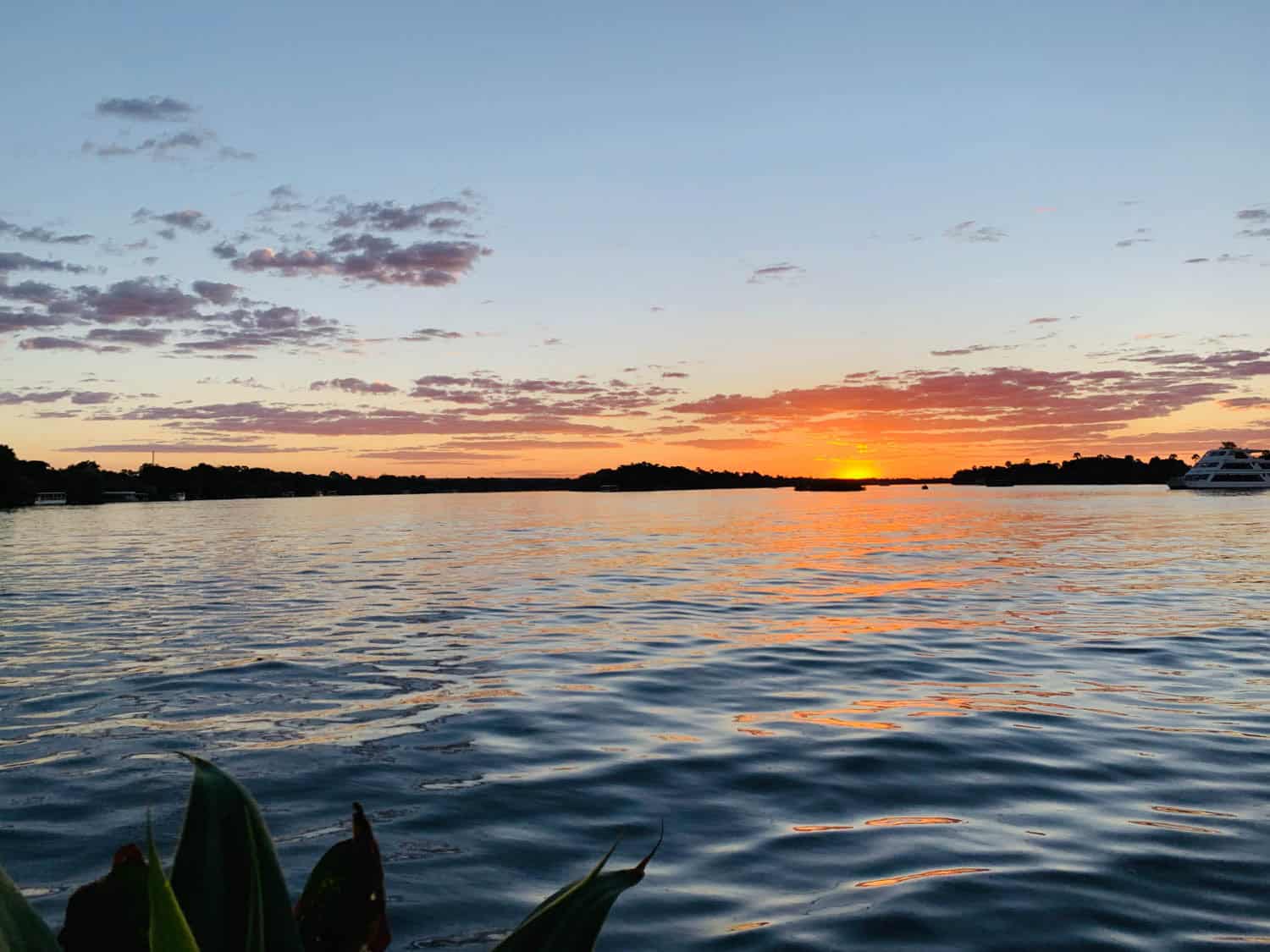 Sunset over a calm lake with orange and pink clouds reflected on the water, shoreline silhouettes in the distance, and a boat on the right side.