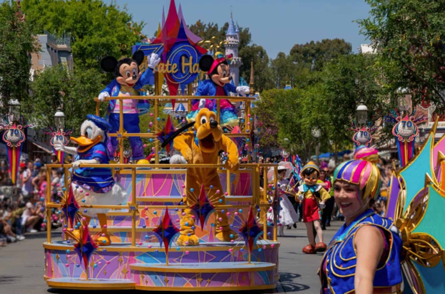 Screenshot A parade float featuring Disney characters Mickey, Minnie, Donald, and Pluto moves down a street, with performers and decorated floats in the background.