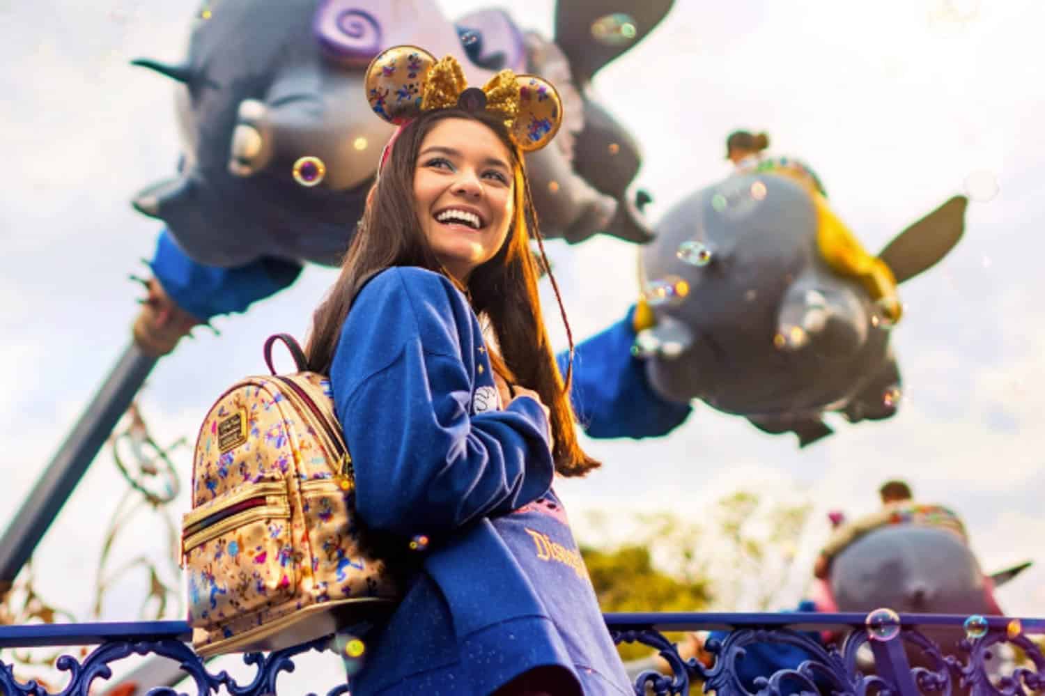Screenshot A young woman wearing Mickey Mouse ears and a backpack smiles at a Disney amusement park, with Dumbo ride attractions and bubbles visible in the background.