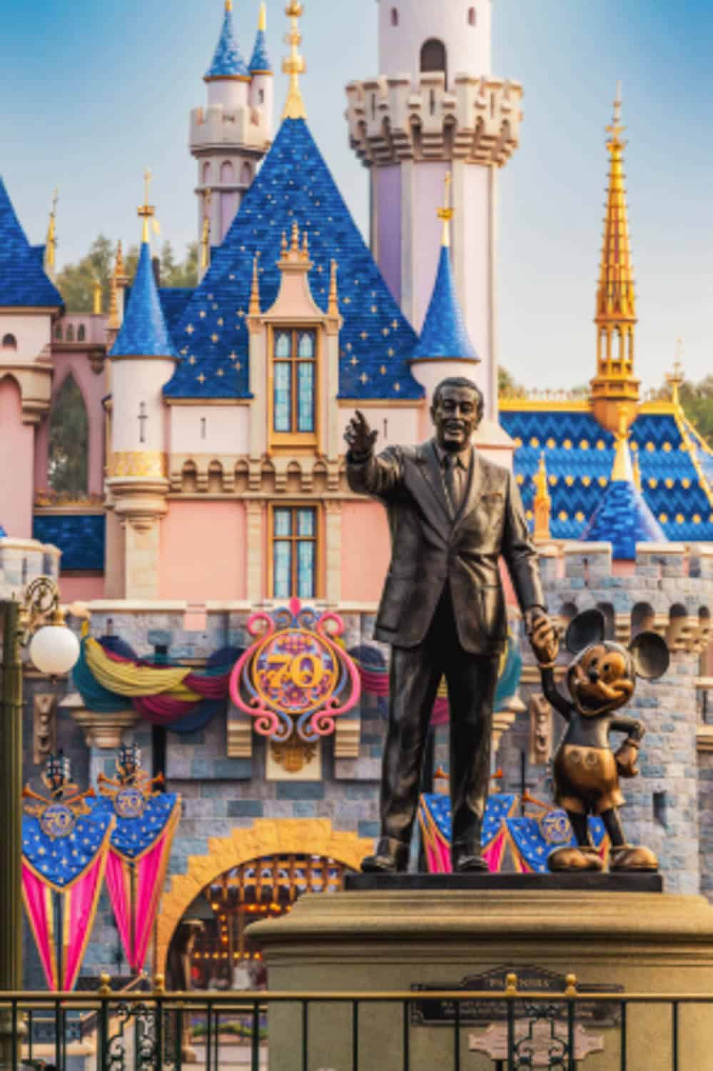 Screenshot Bronze statue of Walt Disney and a Mickey Mouse in front of a colorful castle adorned with blue and gold spires and decorative banners.
