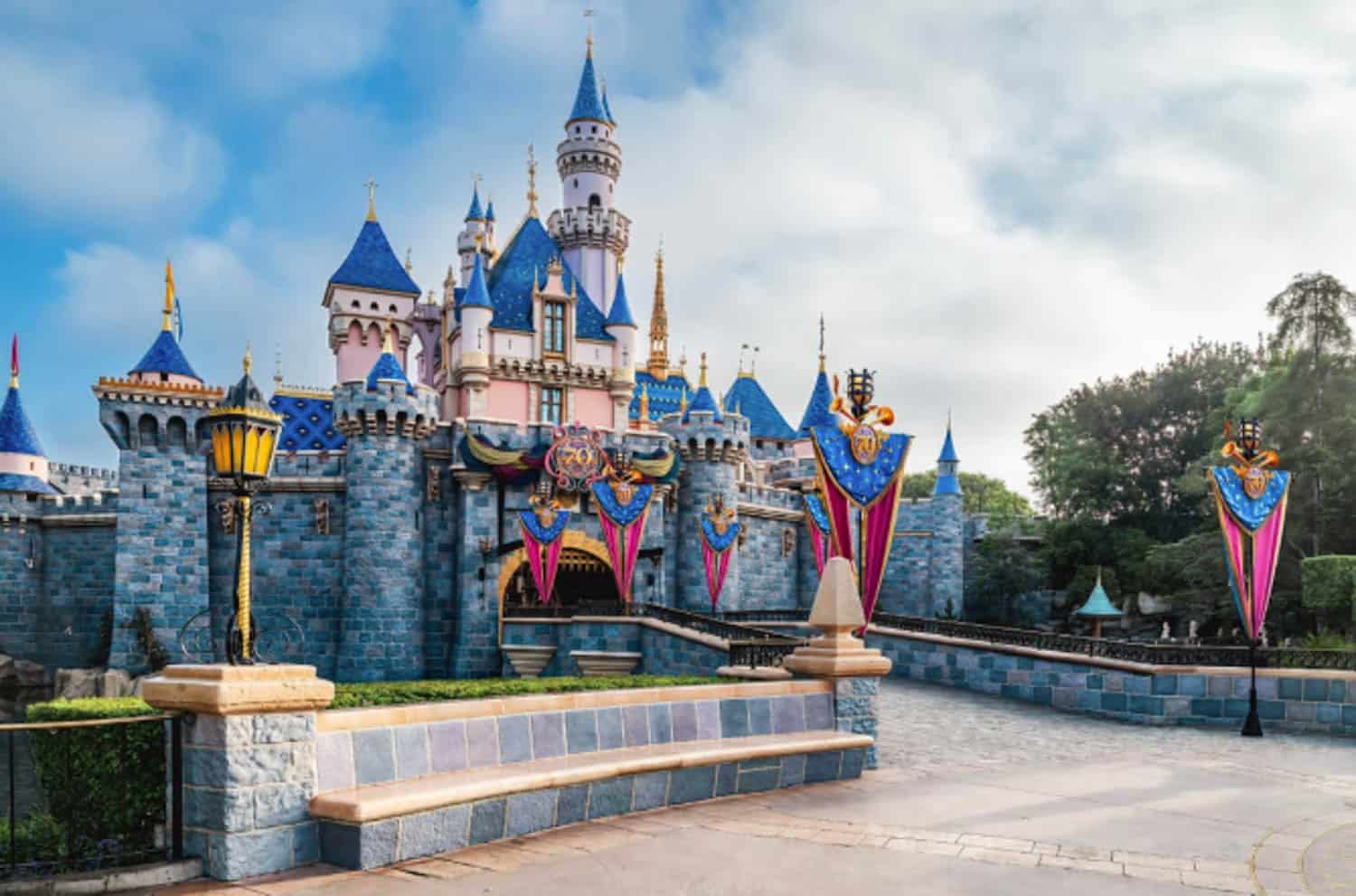Screenshot Sleeping Beauty Castle at Disneyland, decorated with blue and pink banners, surrounded by stone walls and greenery under a partly cloudy sky.