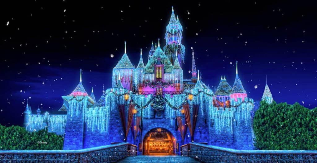 Disneyland castle at night decorated with blue and white holiday lights, with snowflakes falling in the sky.