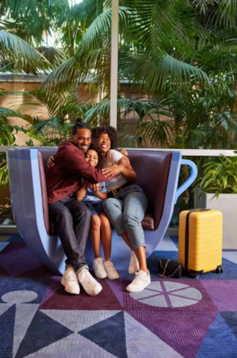 Screenshot A family of three sits closely together in a teacup-shaped chair indoors, smiling, with a yellow suitcase and greenery visible in the background.
