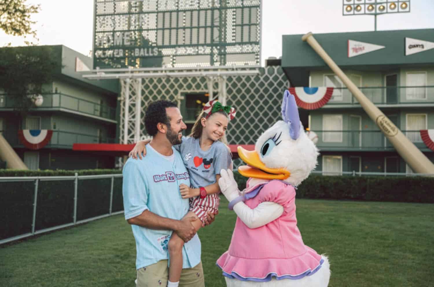Screenshot A man holds a smiling girl as they meet a person in a Daisy Duck costume on a baseball-themed lawn, with a scoreboard and large bats in the background.