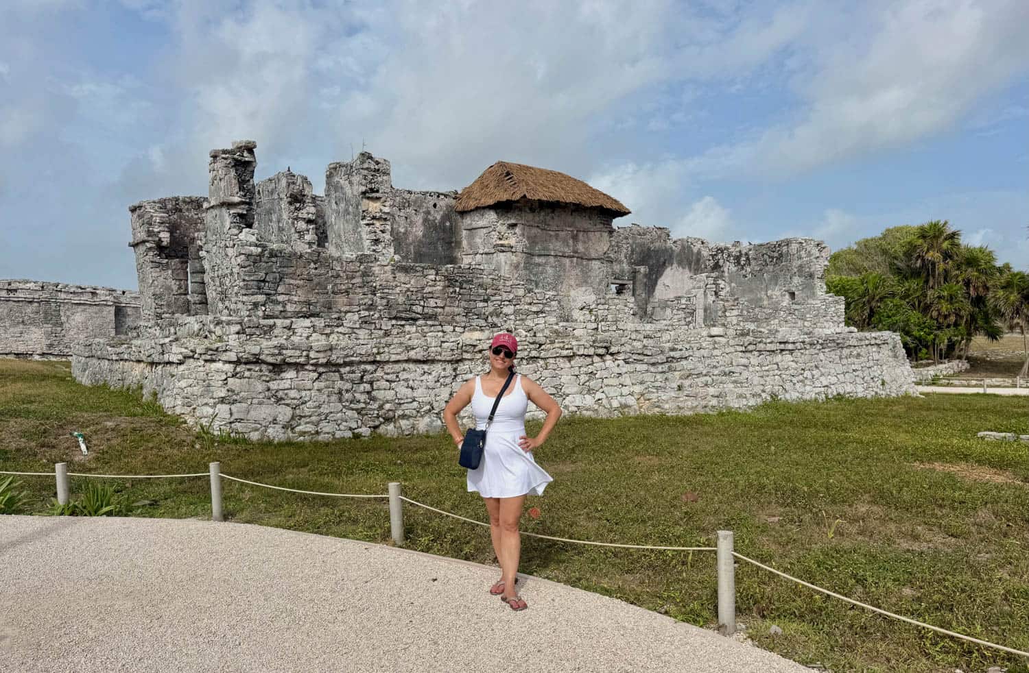 A woman in a white dress and pink cap poses in front of ancient stone ruins under a partly cloudy sky.