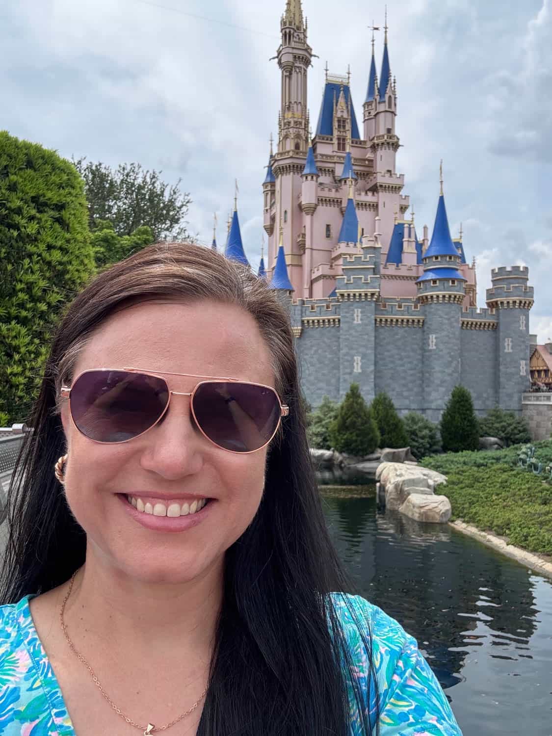 Woman wearing sunglasses smiles in front of Cinderella Castle at a theme park, with a moat, greenery, and blue sky in the background.