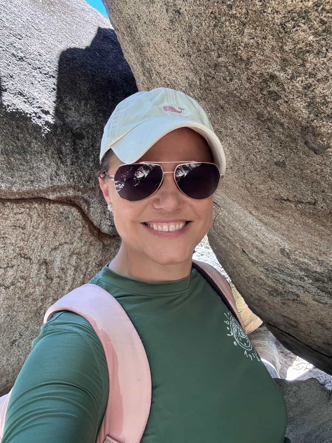 Woman wearing sunglasses, a light-colored cap, and a green shirt smiles for a selfie outdoors between large boulders.