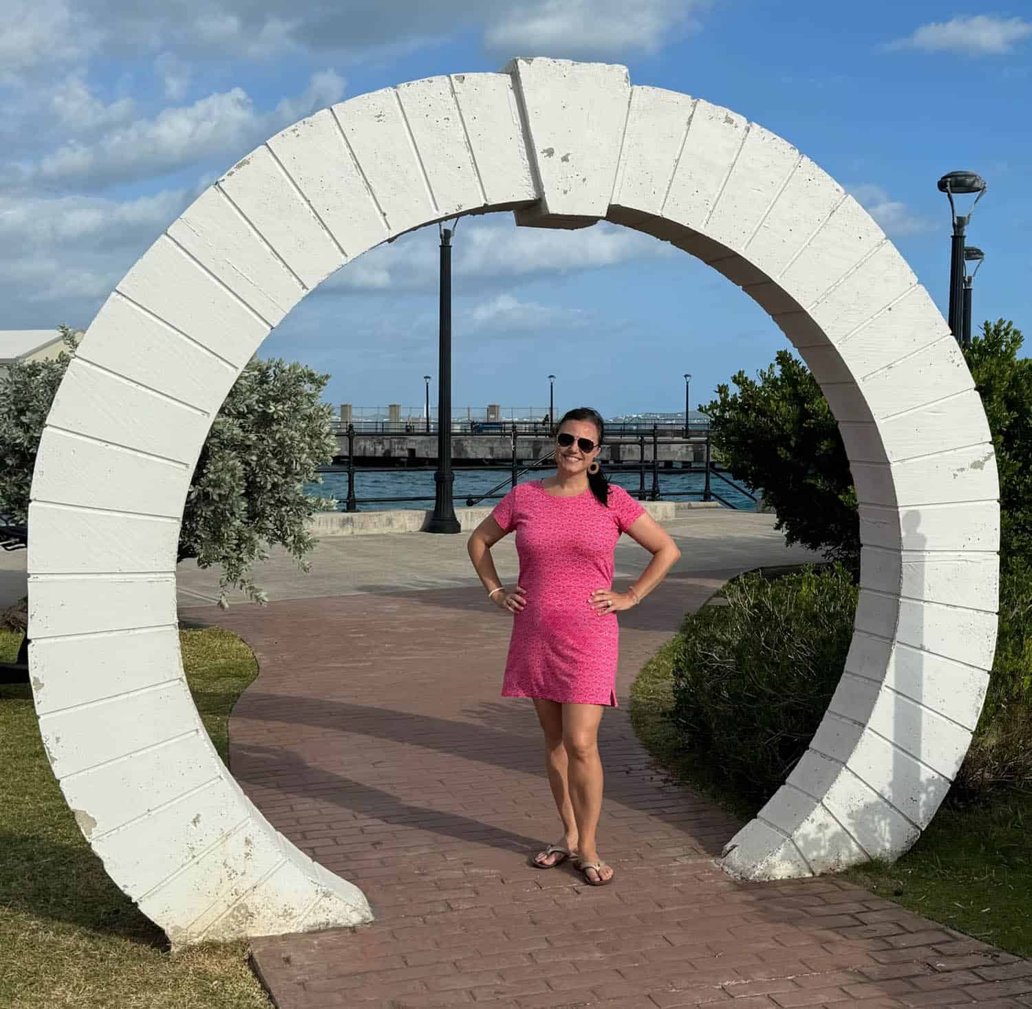 A woman in a pink dress stands smiling under a large, white, circular stone structure by a walkway near water and greenery.