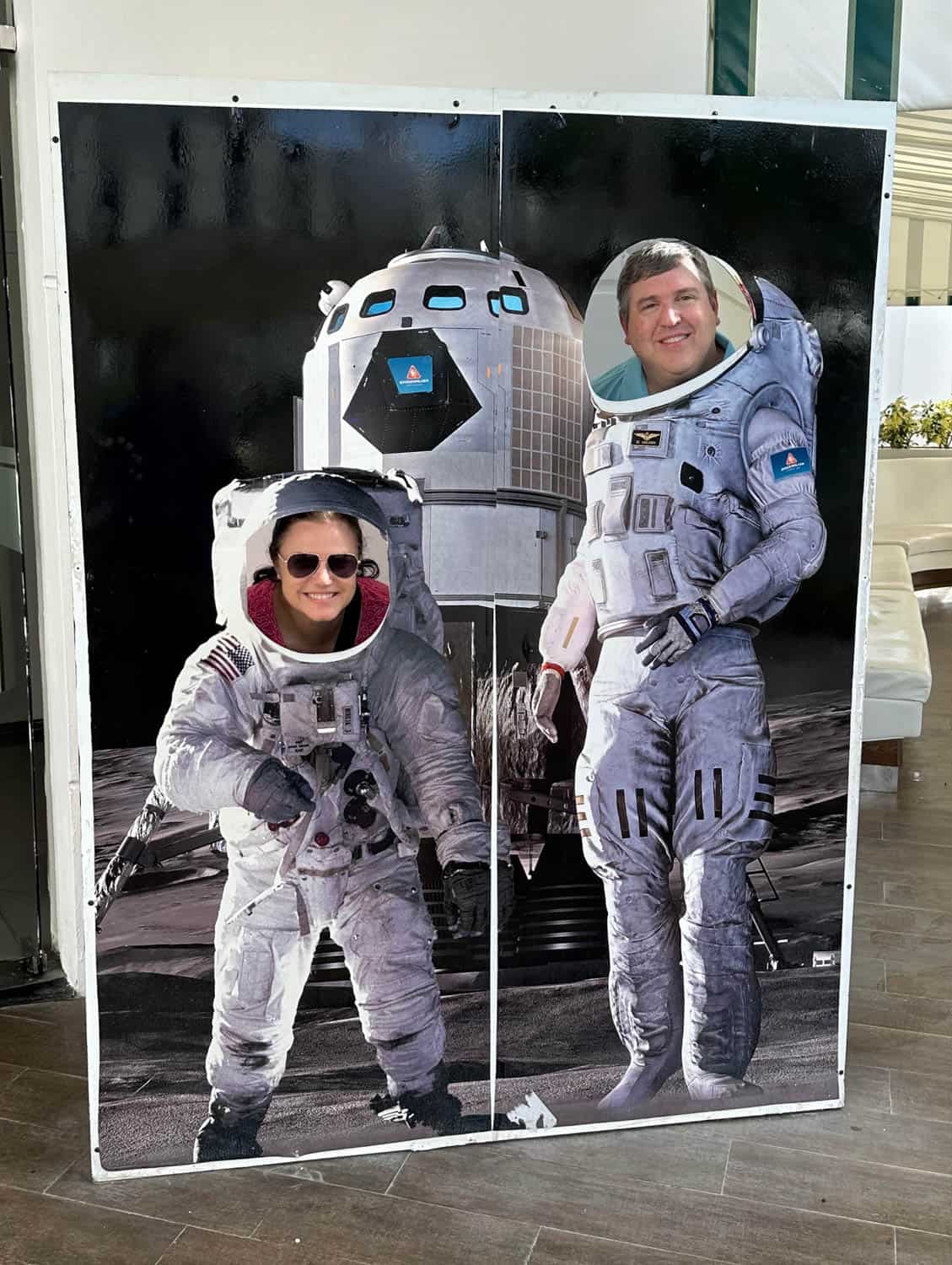 Two people pose behind a cutout board depicting astronauts on the moon, with their faces visible through the face holes of the astronaut suits.