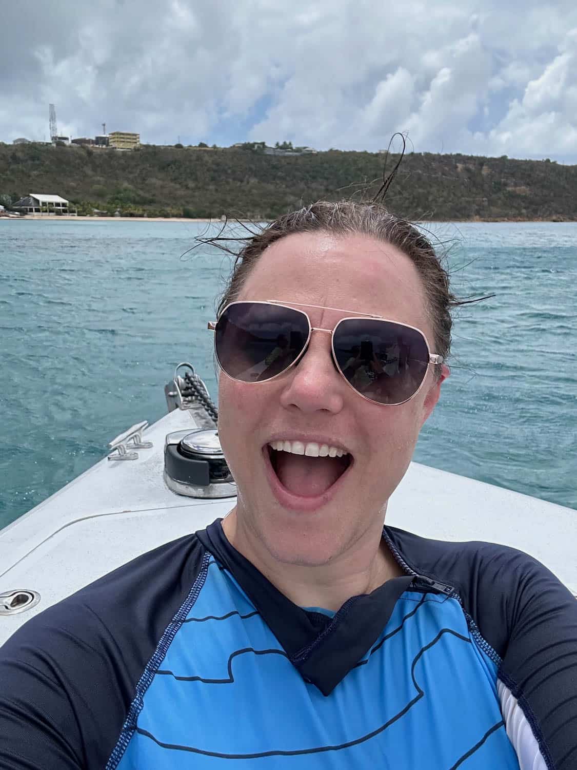 Woman wearing sunglasses and a blue rash guard smiles at the camera while on a boat, with water and a hilly shoreline in the background under cloudy skies.