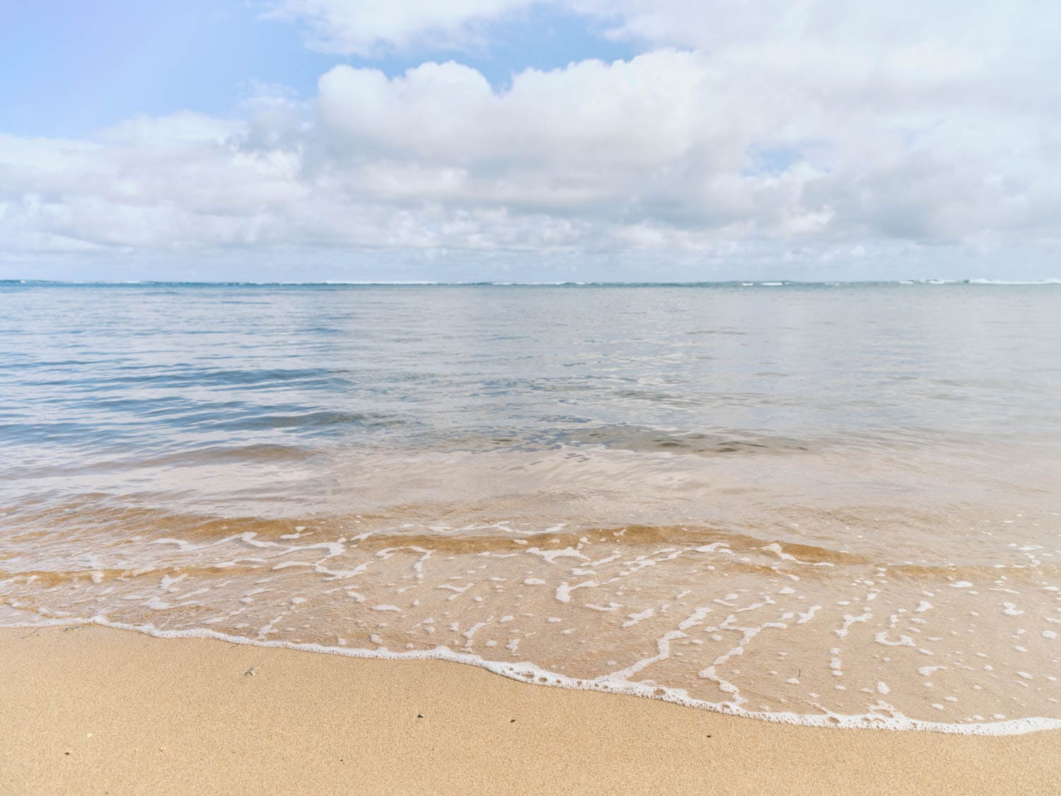 Calm ocean waves gently wash onto a sandy beach under a partly cloudy sky in Hawaii.