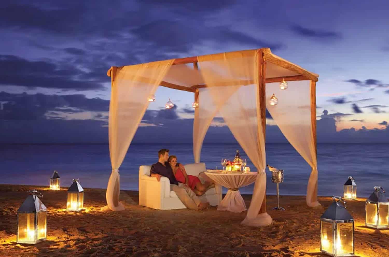 A couple on their honeymoon sits on a white couch under a canopy with hanging lights on a beach at dusk, surrounded by lanterns and a small table set for two.