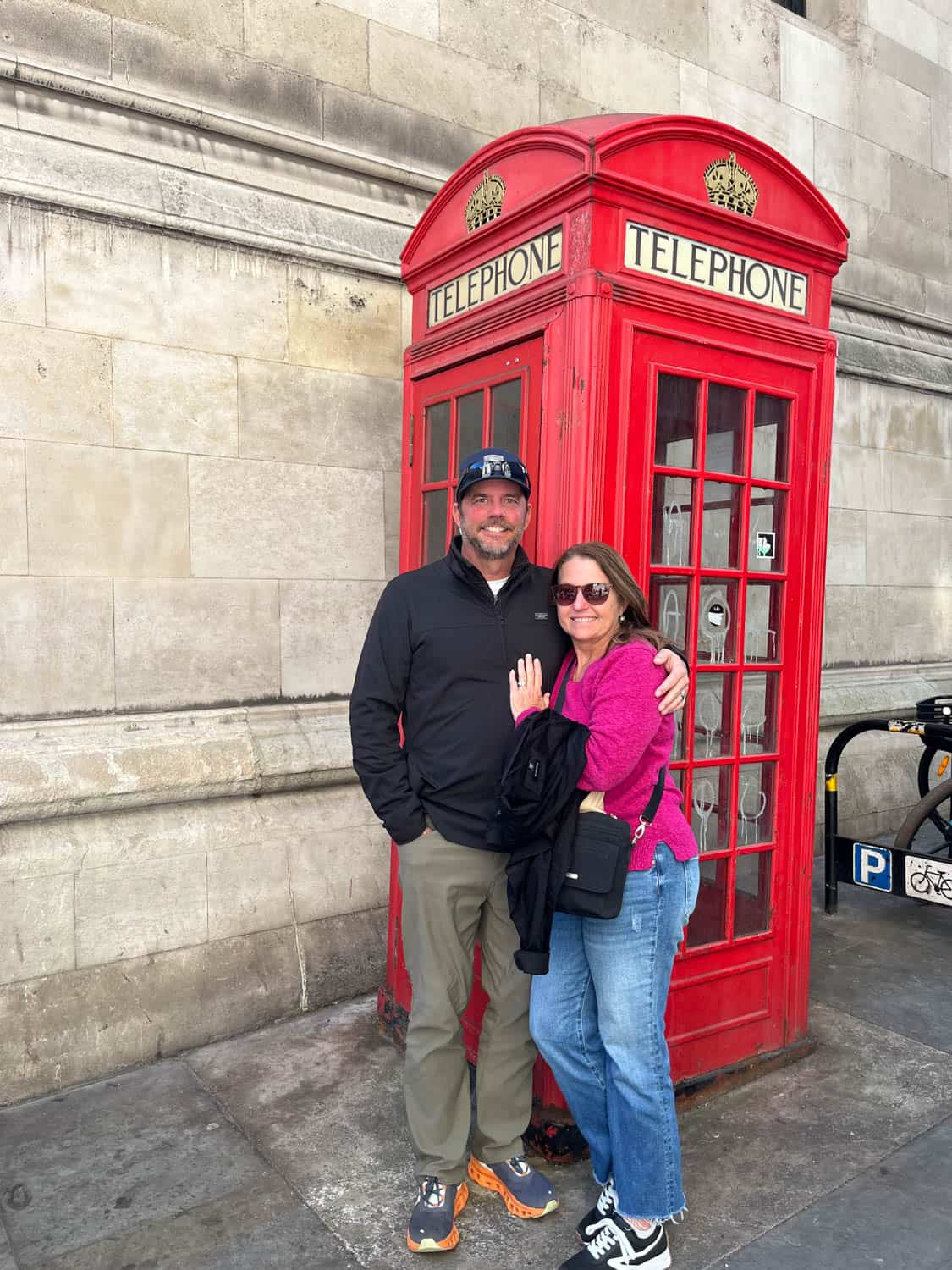 A man and woman stand in front of a classic red British telephone booth on a city sidewalk.
