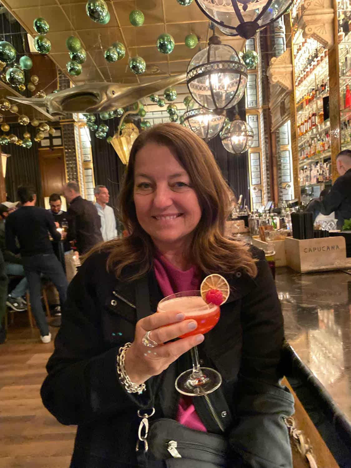 A woman sits at a bar holding a pink cocktail, smiling at the camera. The bar has hanging green ornaments, shelves of bottles, and a mirrored ceiling.