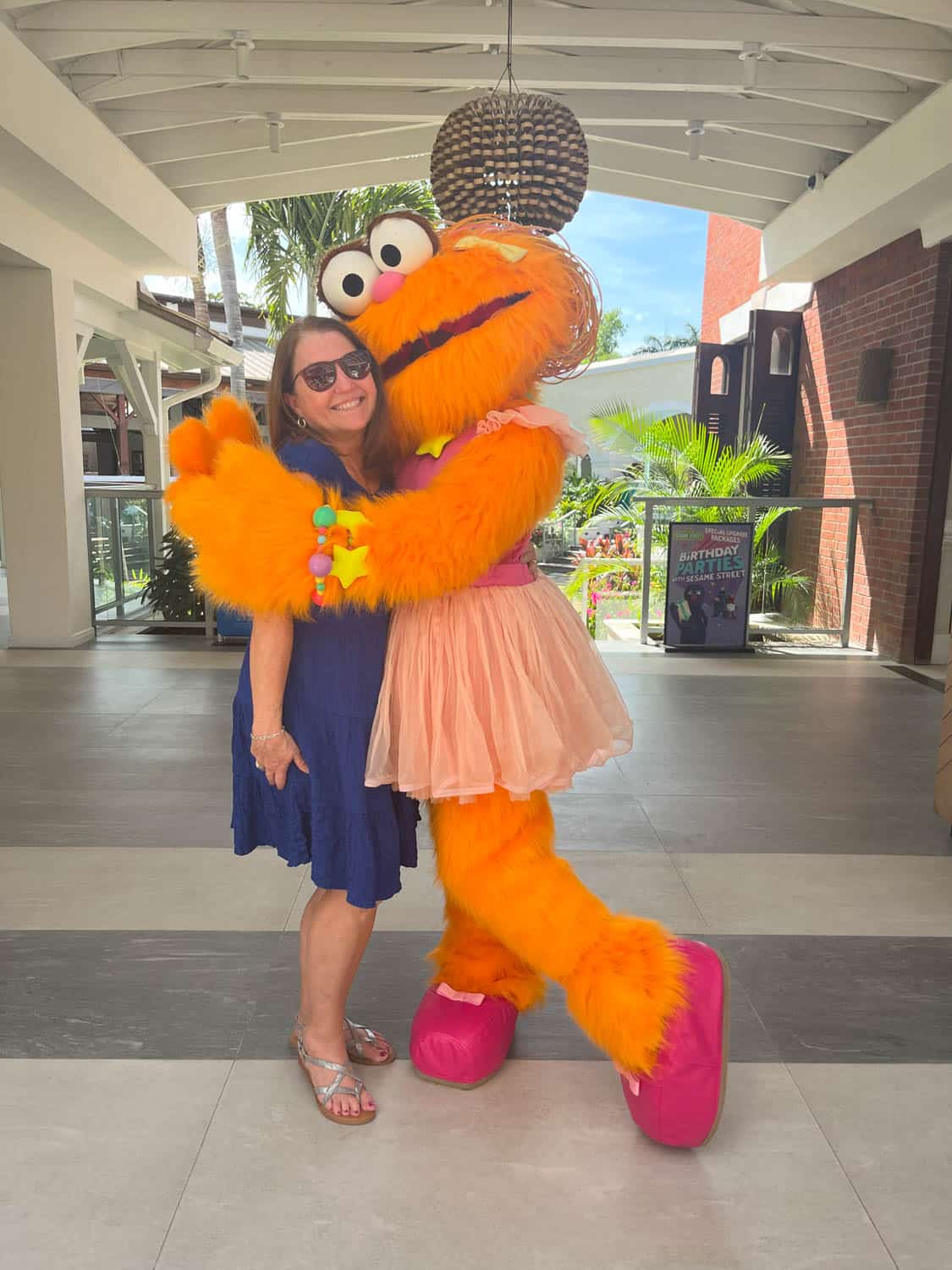 A woman in a blue dress smiles while being hugged by a person in an orange and pink furry character costume indoors.