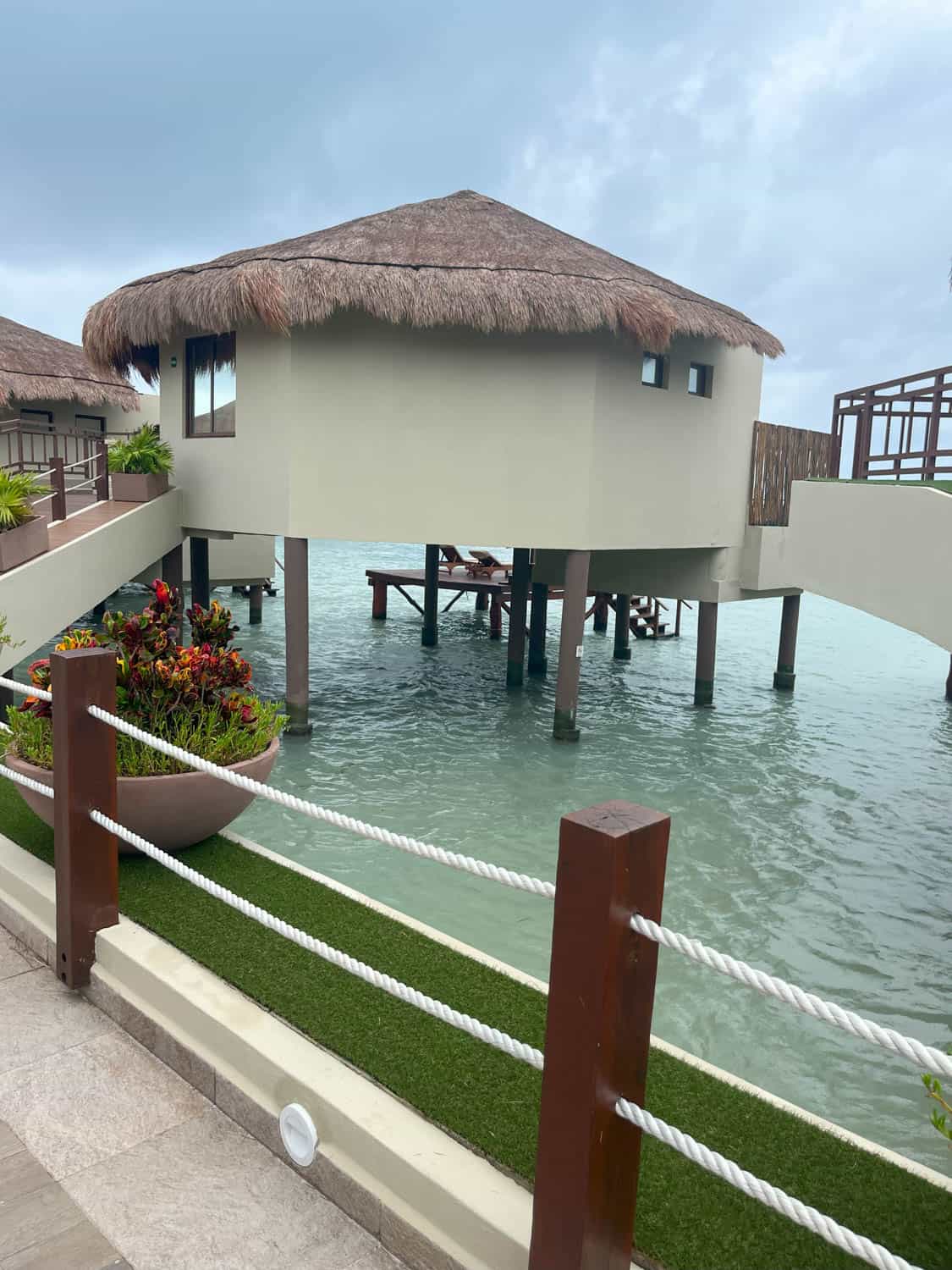 A round overwater bungalow with a thatched roof stands on stilts above clear blue water, surrounded by a wooden walkway and tropical plants.