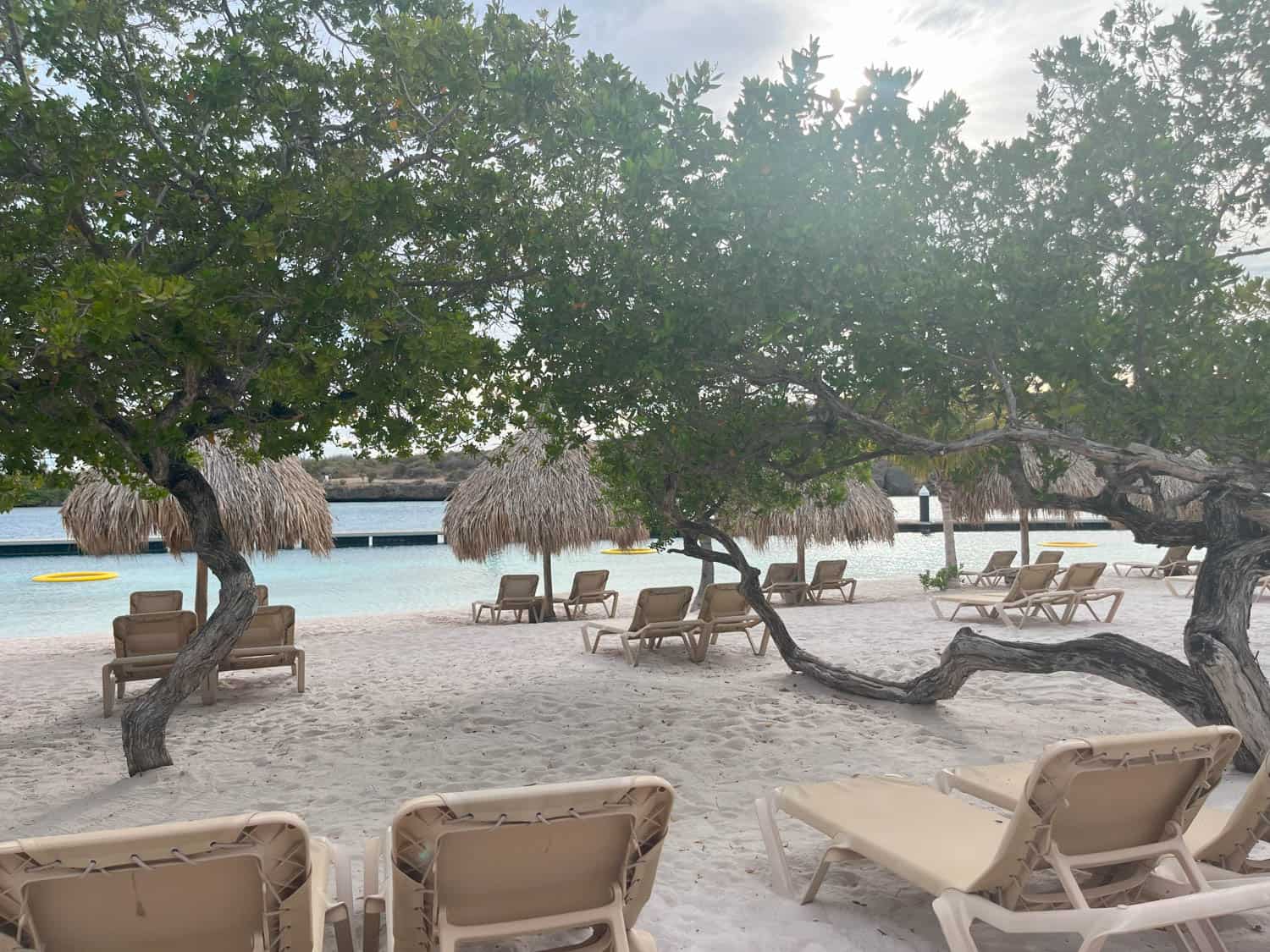 Beach scene with empty lounge chairs on white sand, palm trees, and calm water in the background under a partly cloudy sky.