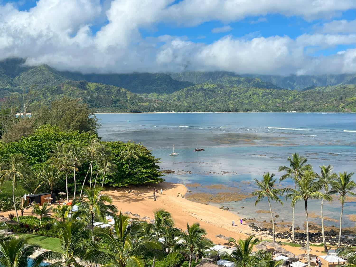 A sandy beach lined with palm trees meets shallow blue water, with green mountains and scattered clouds in the background.
